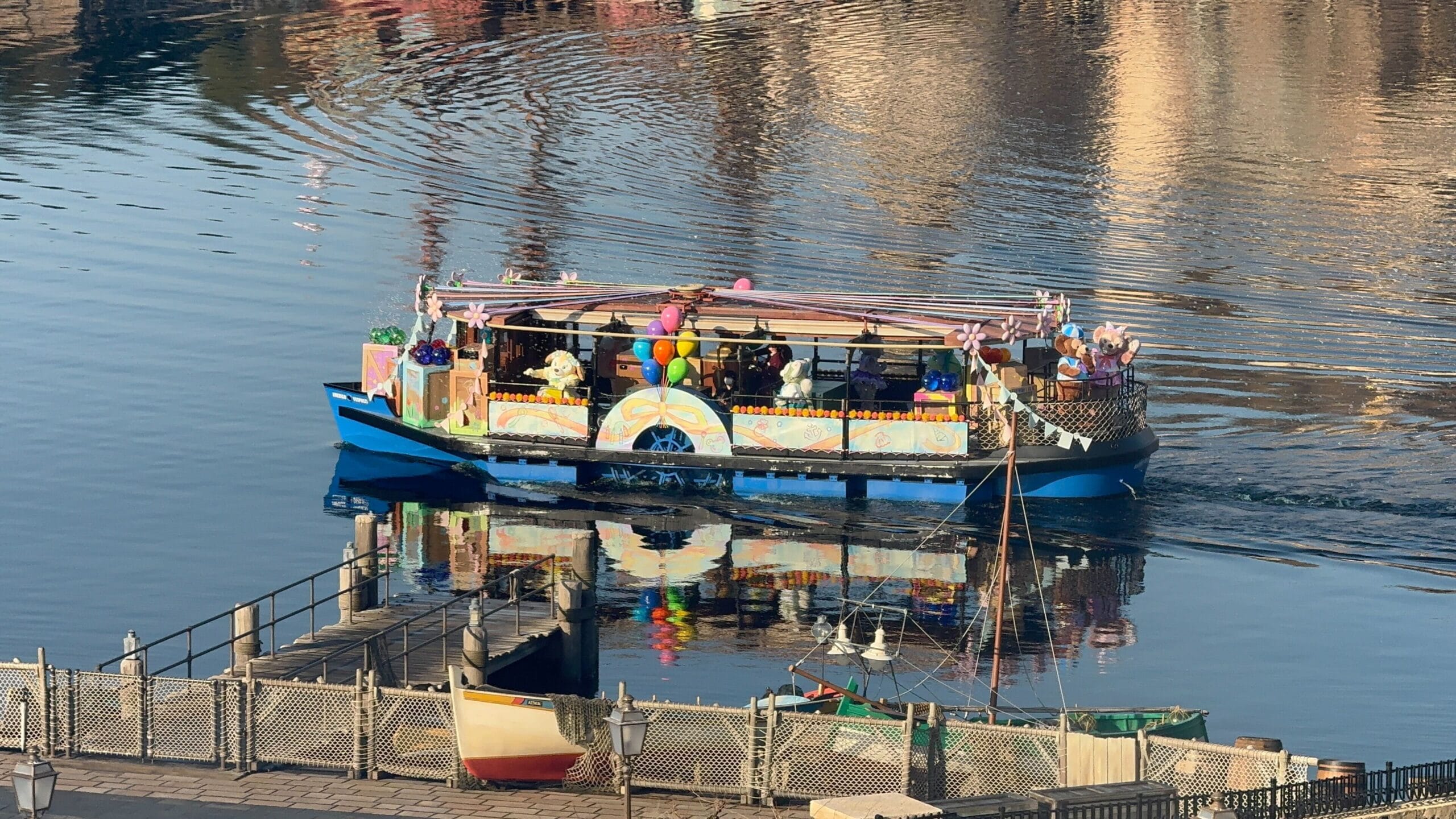 A festive boat decorated with colorful balloons sails on calm water, carrying several passengers.