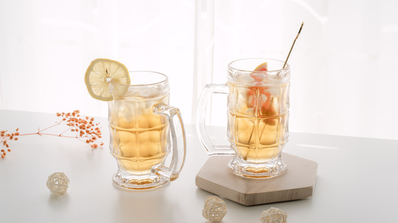 Two steins of pale yellow cocktails; one garnished with a lemon wheel and the other with a slice of grapefruit.