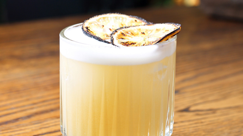 A pale yellow cocktail in a rocks glass with egg foam on top. Garnished with toasted lemon wheels.