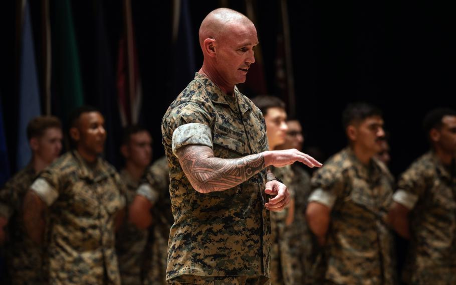 A U.S. Marine in camouflage uniform stands in the foreground, gesturing with his right hand while speaking. He has a shaved head and visible sleeve tattoo on his right arm. Behind him, a formation of fellow Marines stands in rows, also wearing camouflage uniforms. The lighting highlights the speaker while the others remain slightly out of focus.