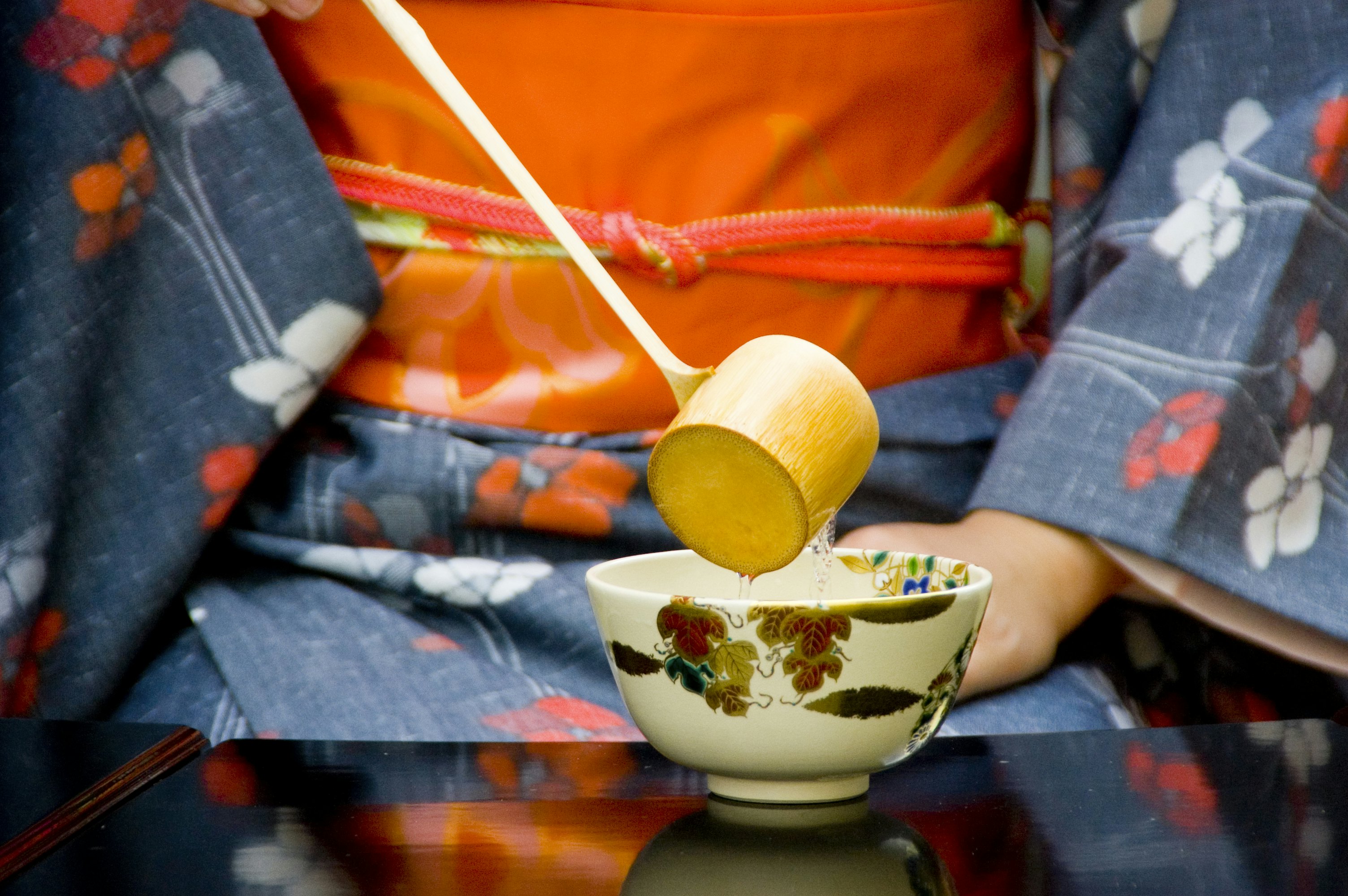 A person conducting a tea ceremony, wearing a blue kimono and orange obi pouring hot water from a wooden ladle into a Japanese tea cup