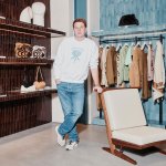 Portrait of Jonathan Anderson in a Loewe retail store. He wears a crewneck sweater and light-wash jeans and leans on a wood chair.