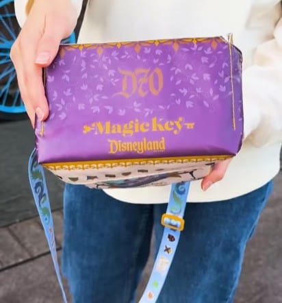 Person holding a purple Disneyland Magic Key insulated cooler bag with gold lettering and a blue character strap.