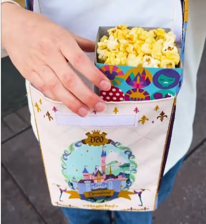 A person holds a Magic Key Disneyland popcorn holder worn around their neck, filled with popcorn.