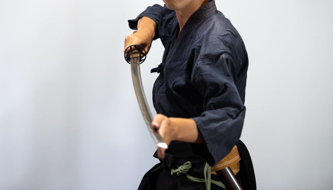 Man holding Japanese sword pointed at viewer with left hand resting against edge near the point