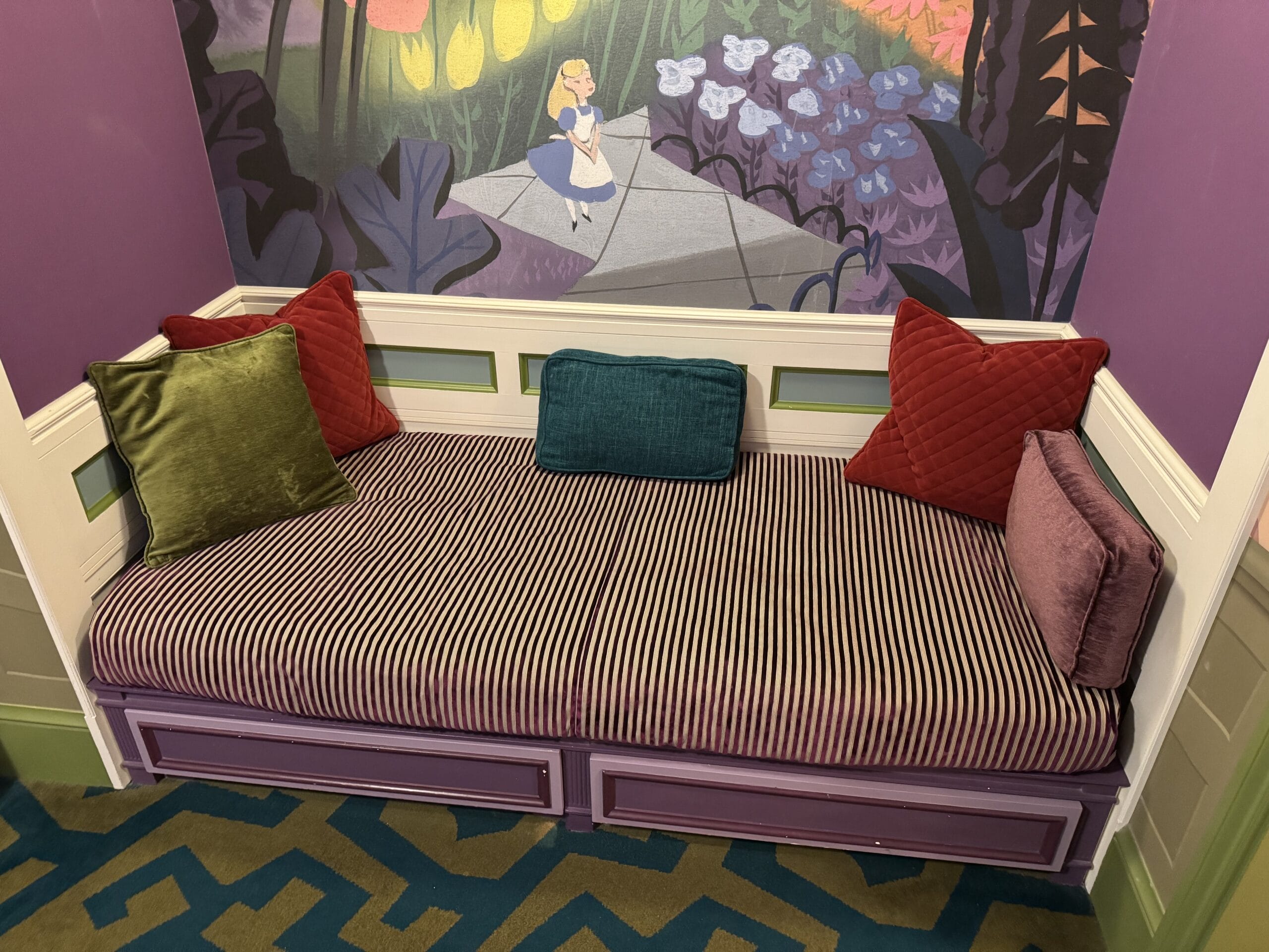 A striped purple daybed with colorful pillows sits beneath an Alice in Wonderland mural at Tokyo Disneyland Hotel.