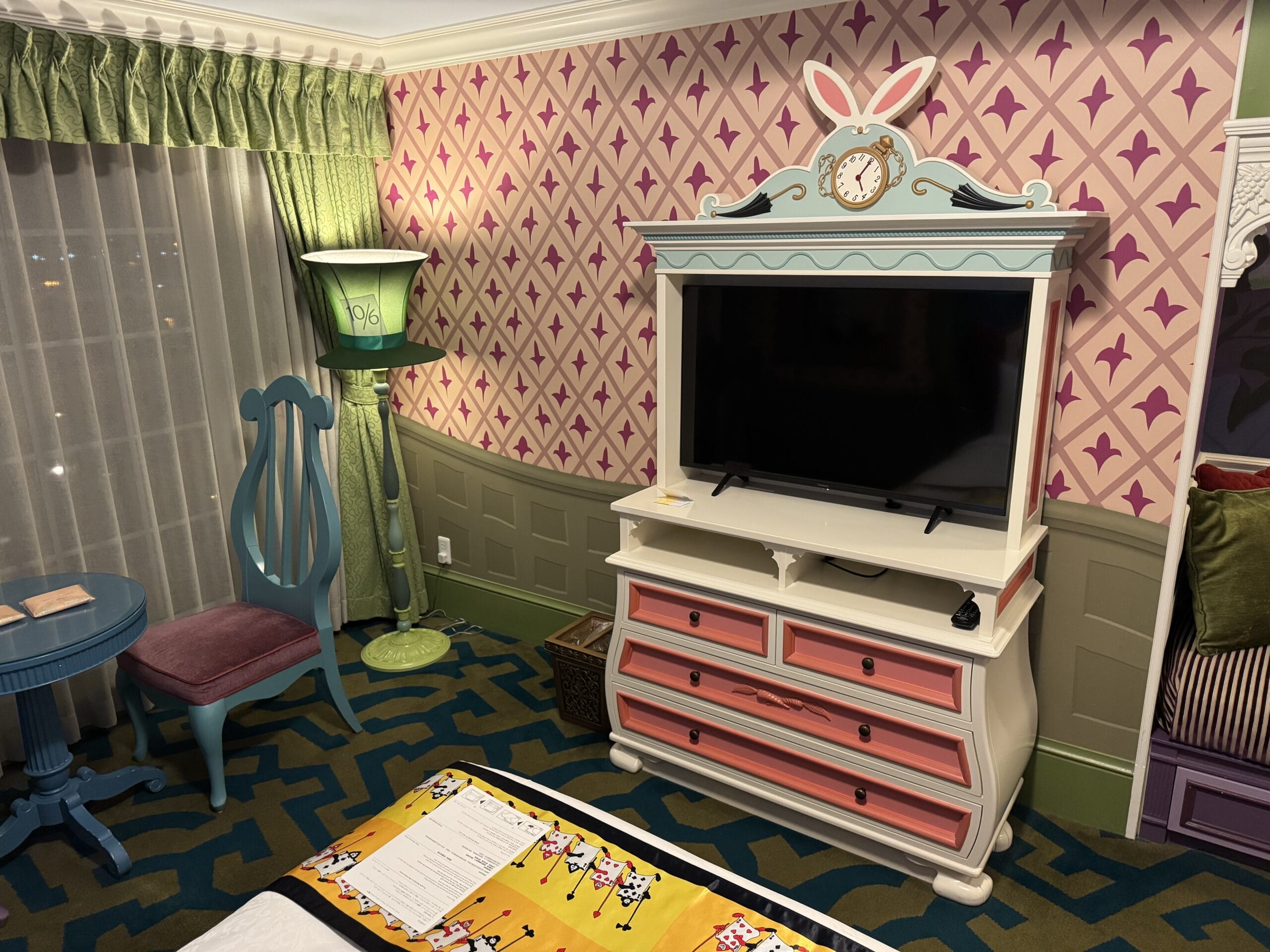 Whimsical Alice in Wonderland hotel room with bunny TV cabinet, pink wallpaper, green curtains, and blue furniture.