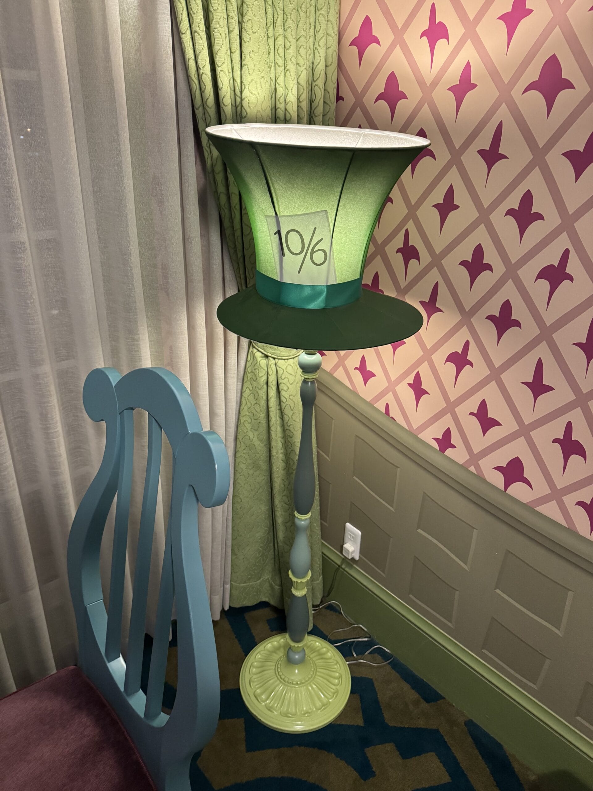 A floor lamp with a Mad Hatter's hat shade stands by a teal chair in this Alice in Wonderland hotel room.