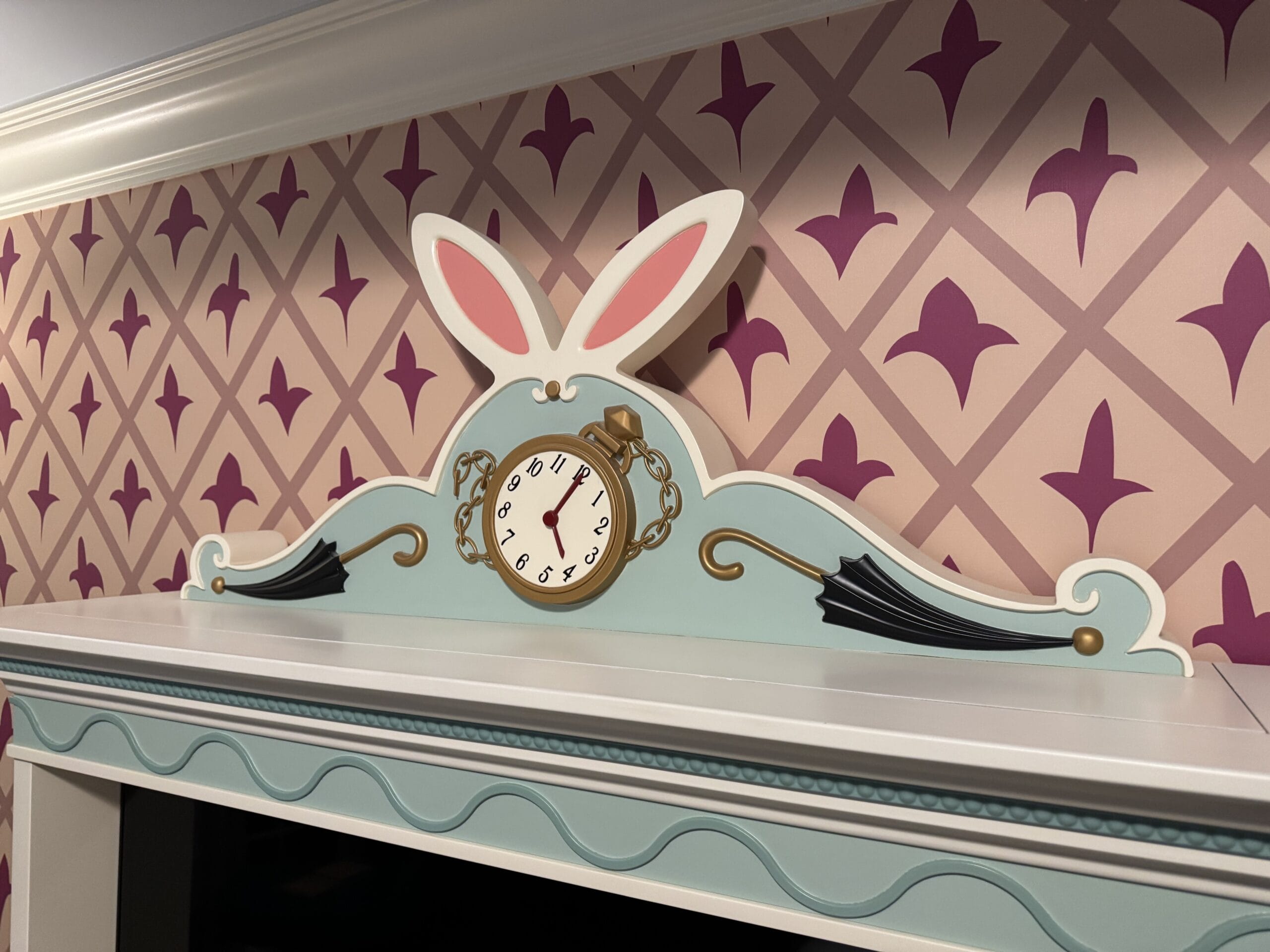 Pastel clock with rabbit ears sits on a mantel in an Alice in Wonderland hotel room tour against pink geometric wallpaper.