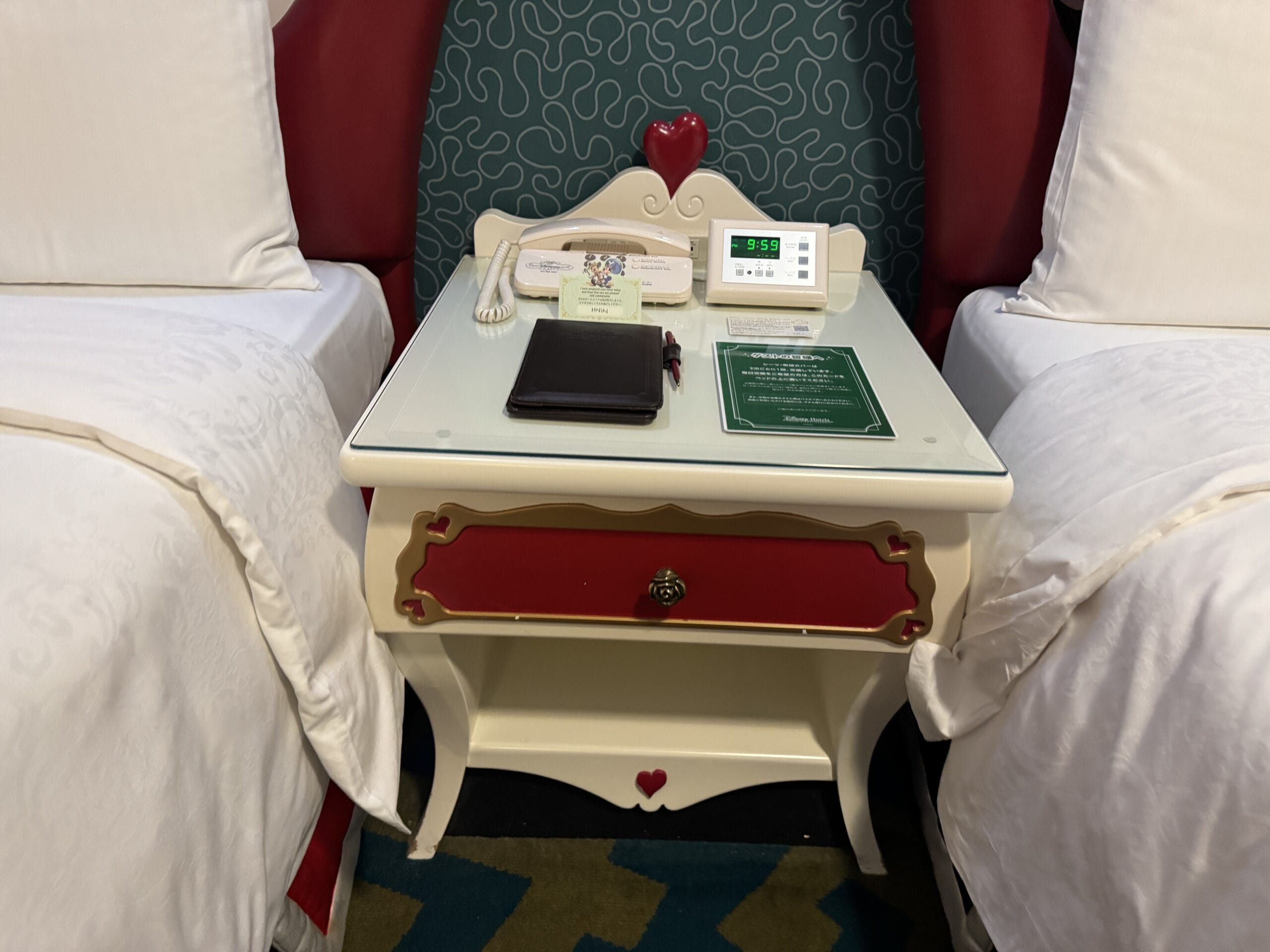 A nightstand with a clock, phone, and notepad sits between two white beds in an Alice in Wonderland Hotel room.