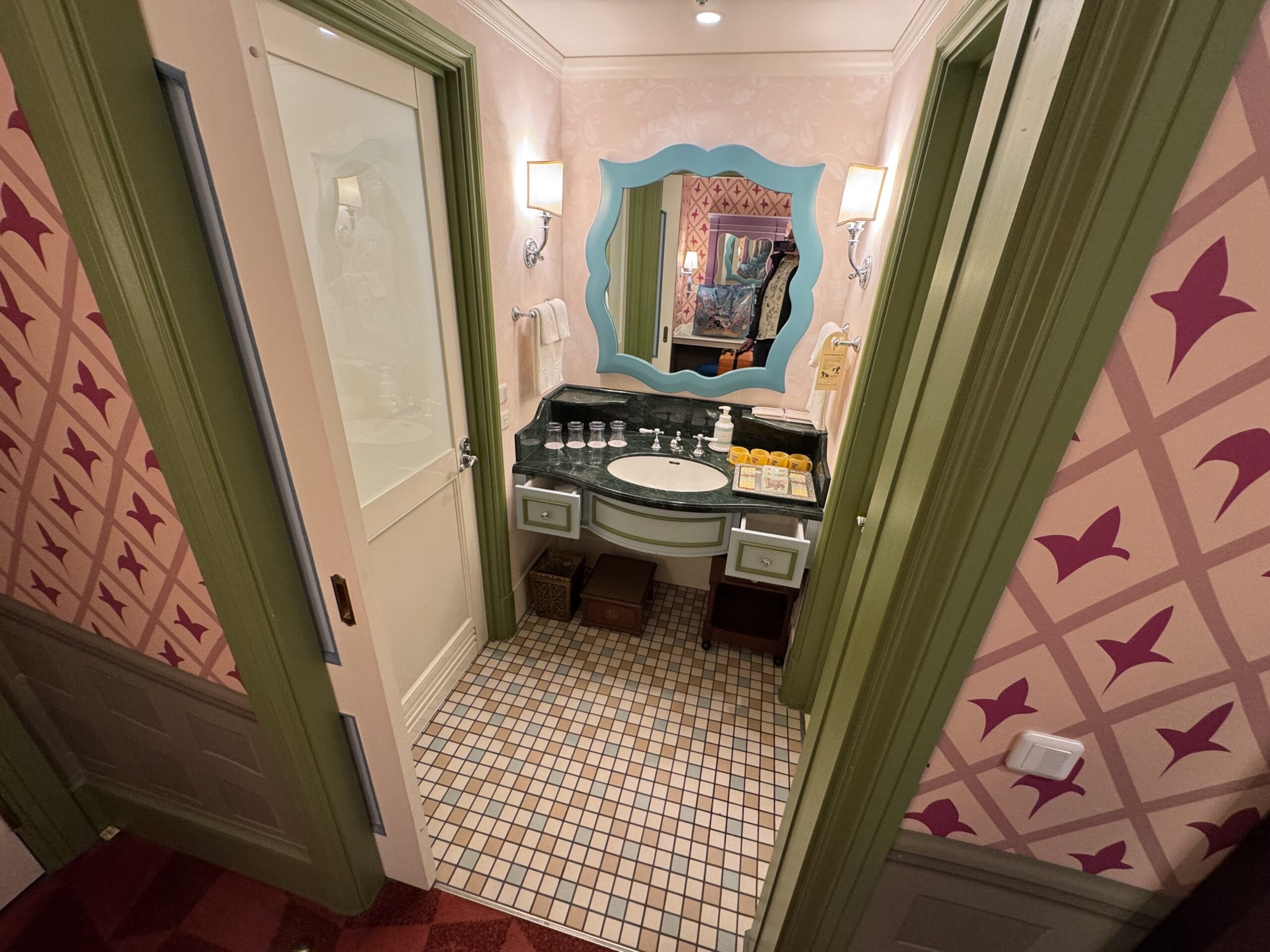 A bathroom at the Tokyo Disneyland Hotel with a green vanity, large mirror, and pink Alice in Wonderland wallpaper.