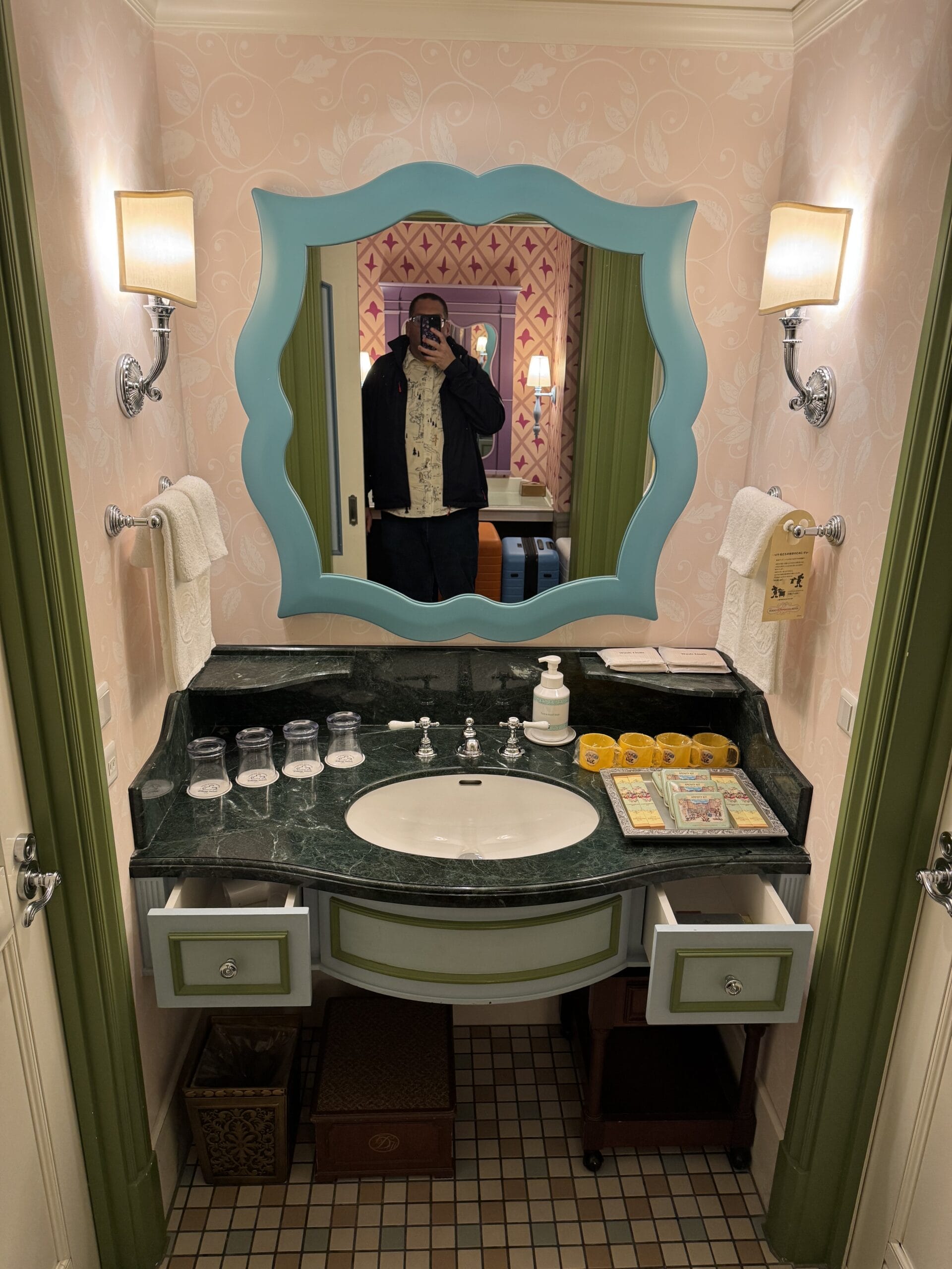 A person takes a mirror selfie in a vintage-style Alice in Wonderland hotel bathroom with green accents and open drawers.