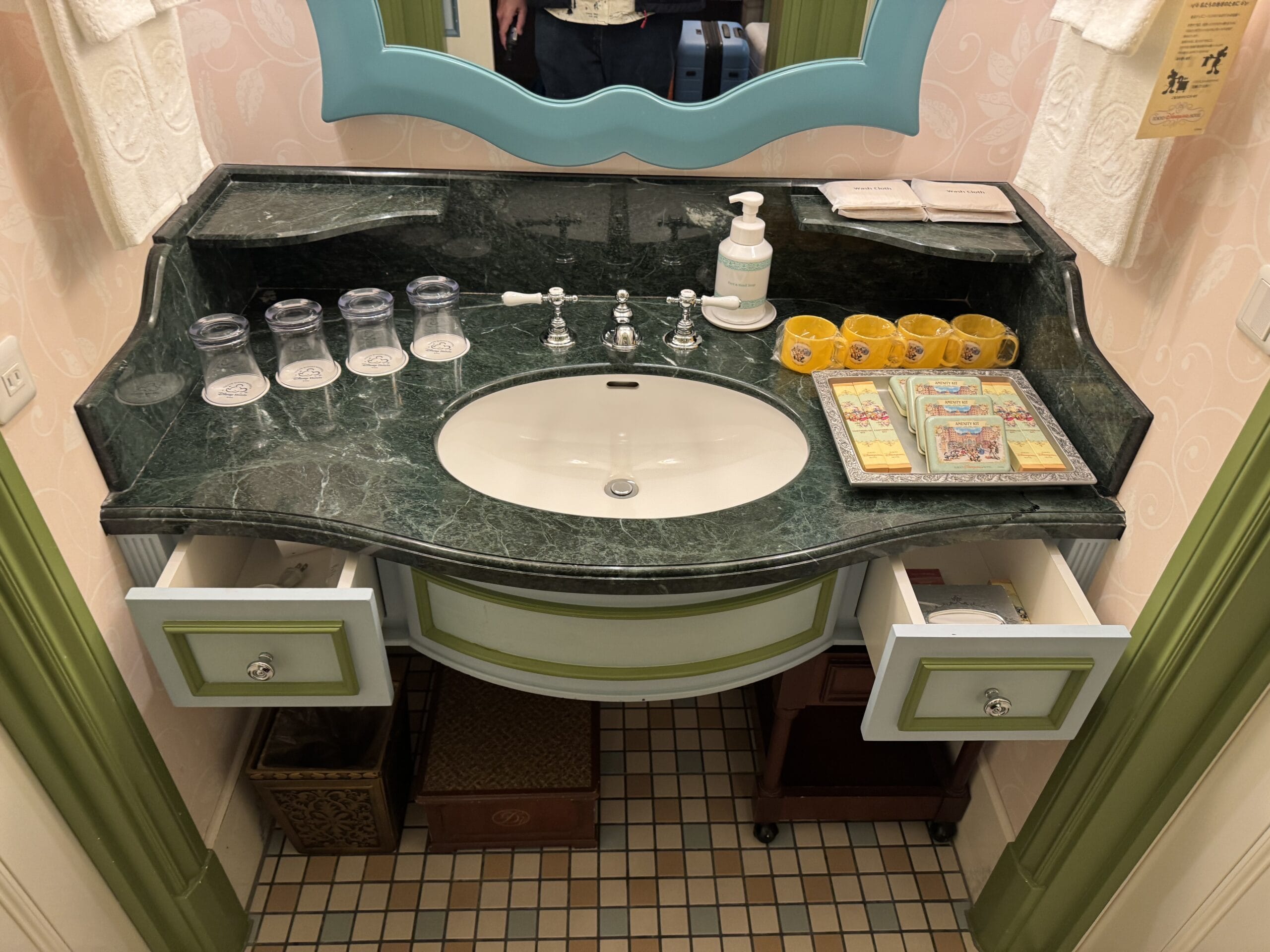 A bathroom sink in an Alice in Wonderland hotel room with amenities, mugs, and open drawers at Tokyo Disneyland Hotel.