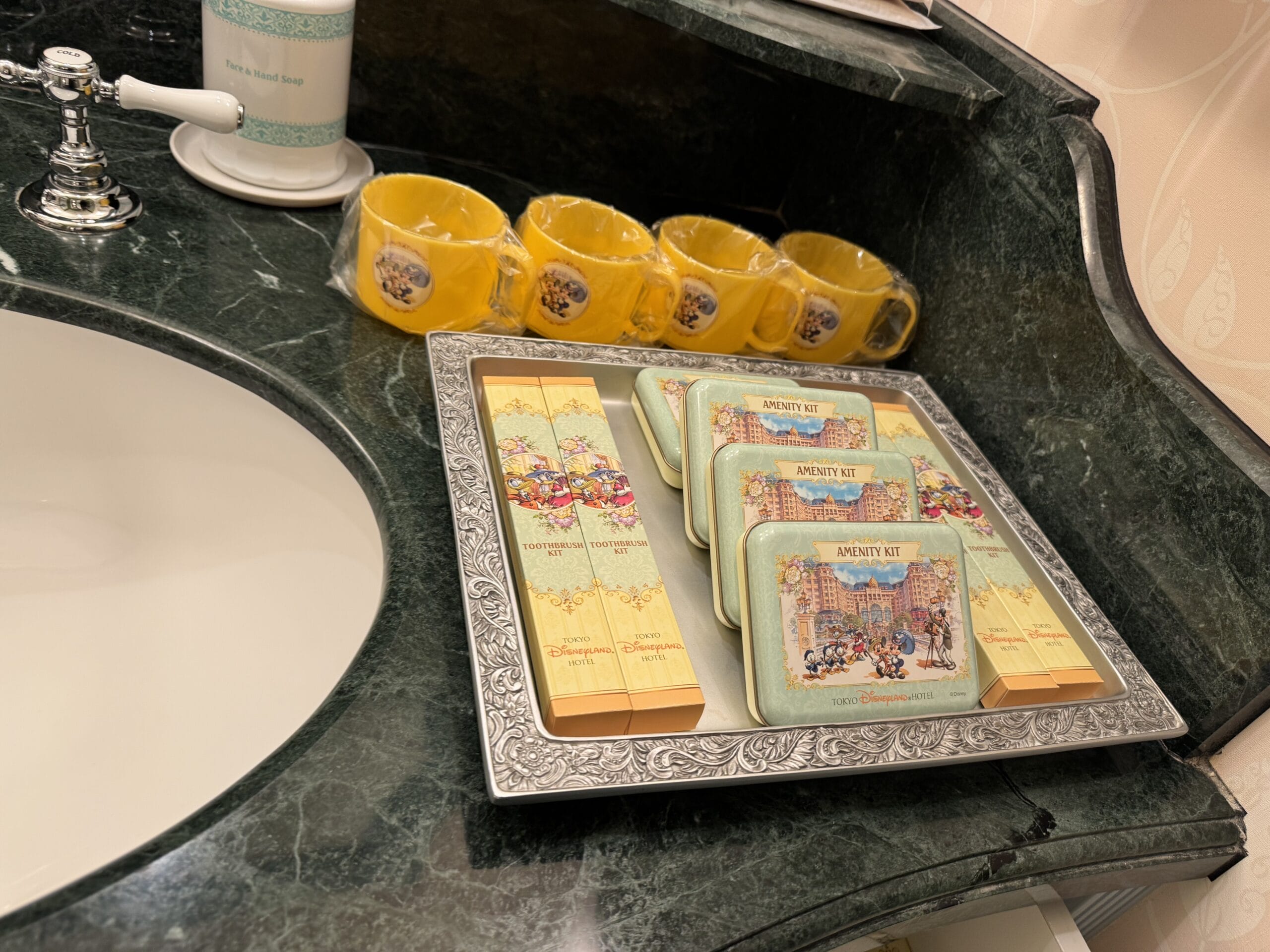 A silver tray with Alice in Wonderland hotel toiletry kits and cups sits on a dark green bathroom counter by the sink.