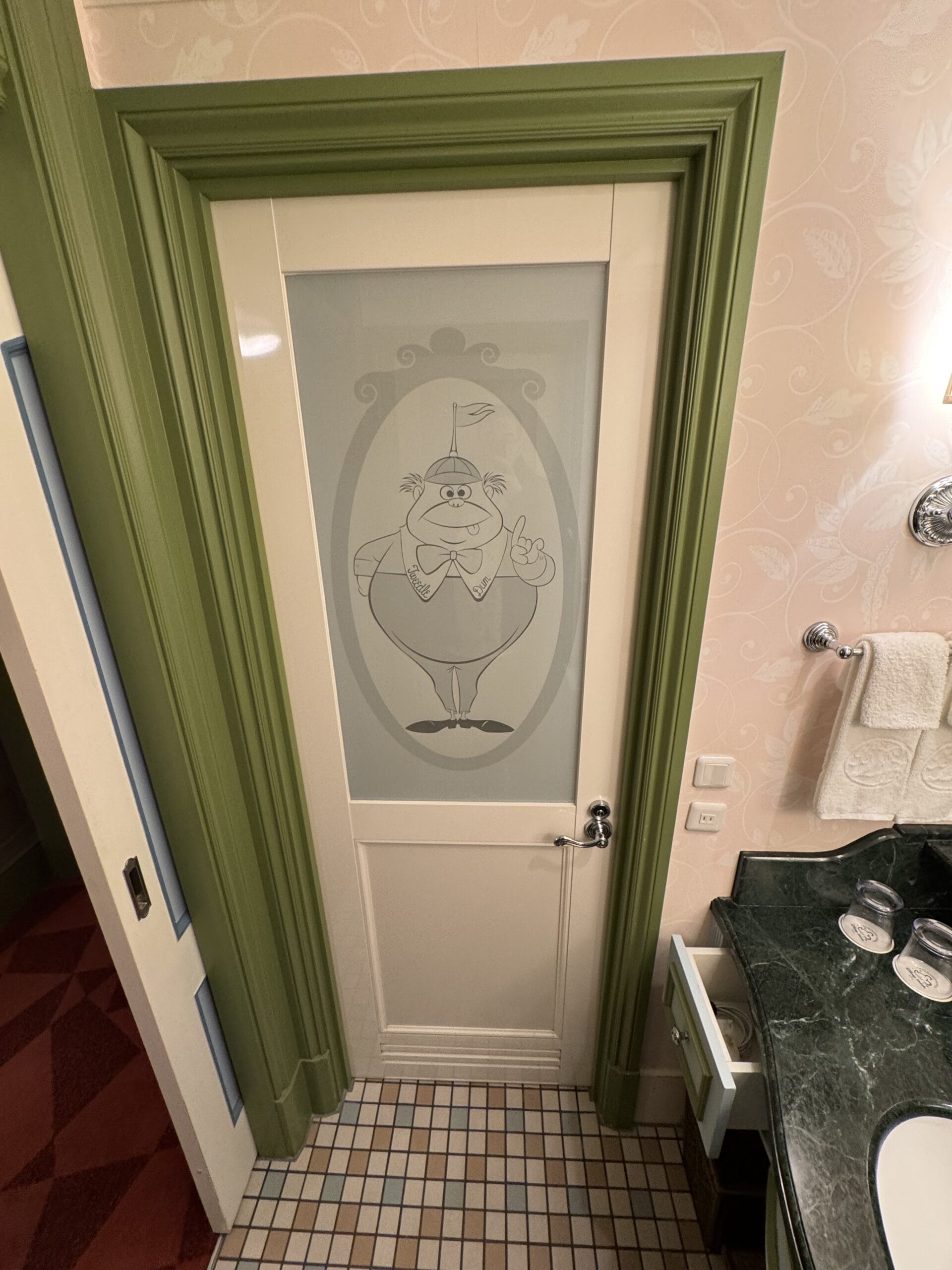 Bathroom door at the Alice in Wonderland Hotel, with a cartoon character etched on frosted glass, beside the sink and towels.