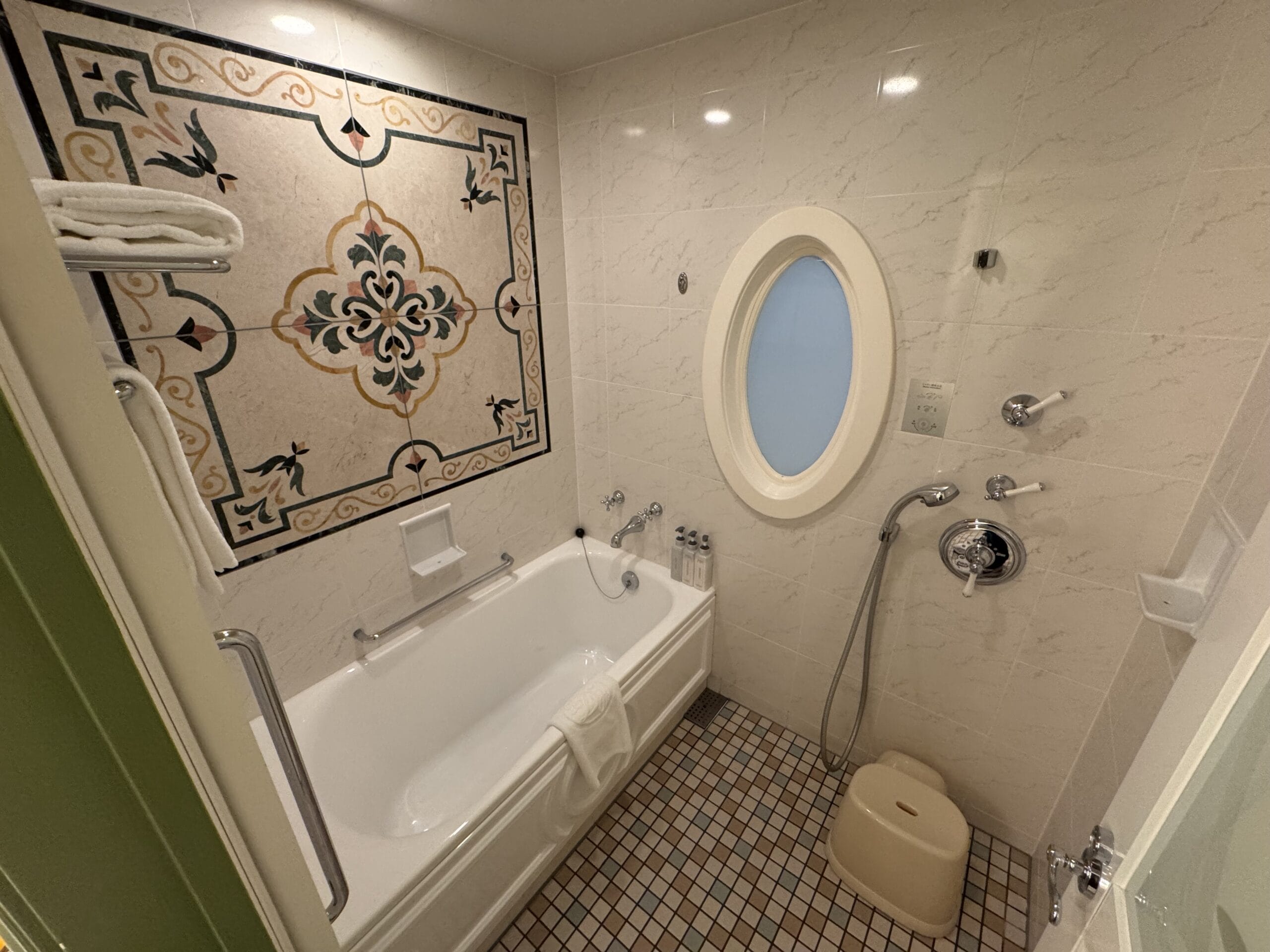 A whimsical bathroom at the Tokyo Disneyland Hotel features a tub, tiled wall, oval window, and handheld shower.