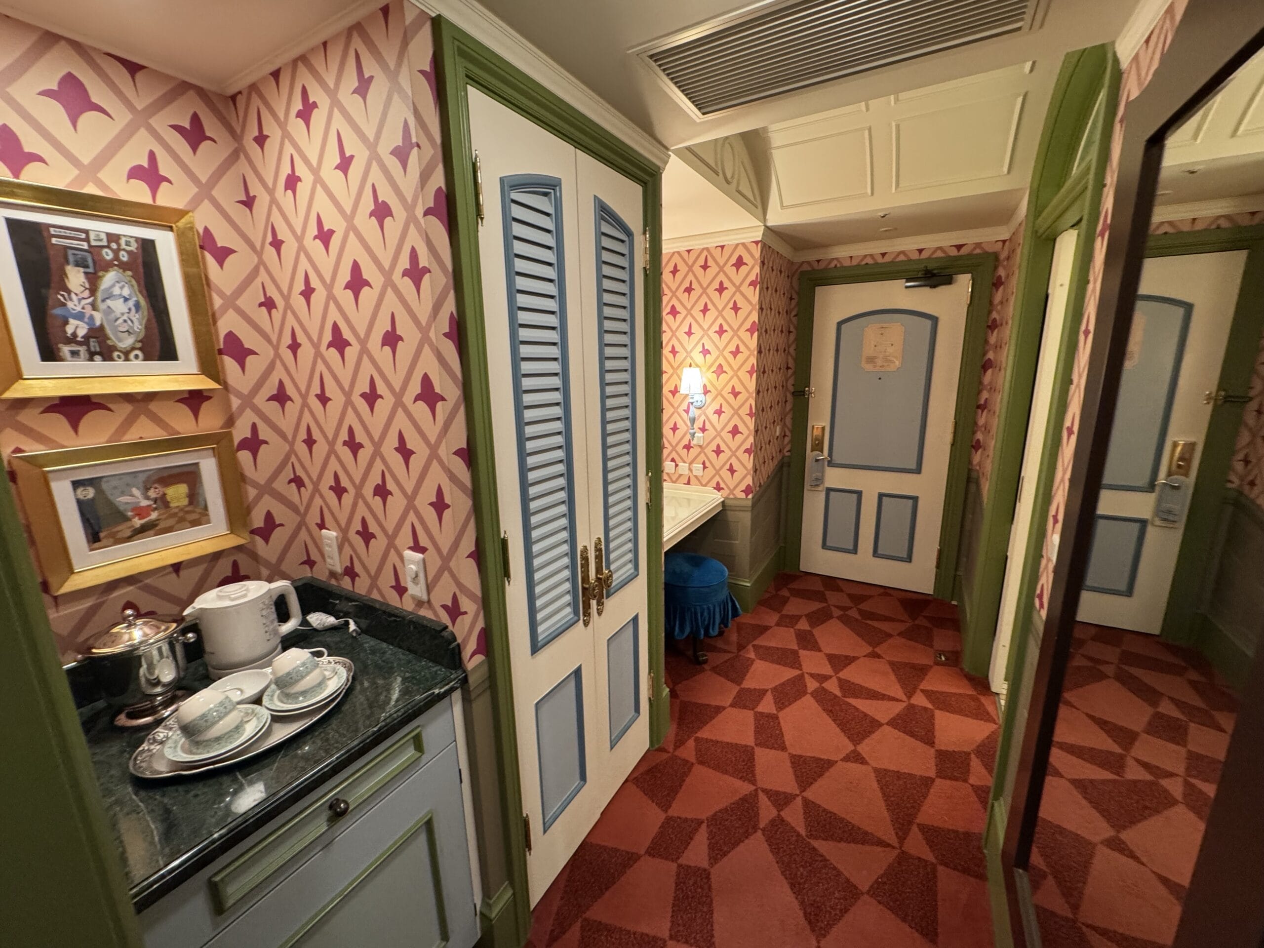 A patterned Alice in Wonderland Hotel Room with a tea set, art, blue doors, mirror, and pink geometric wallpaper.