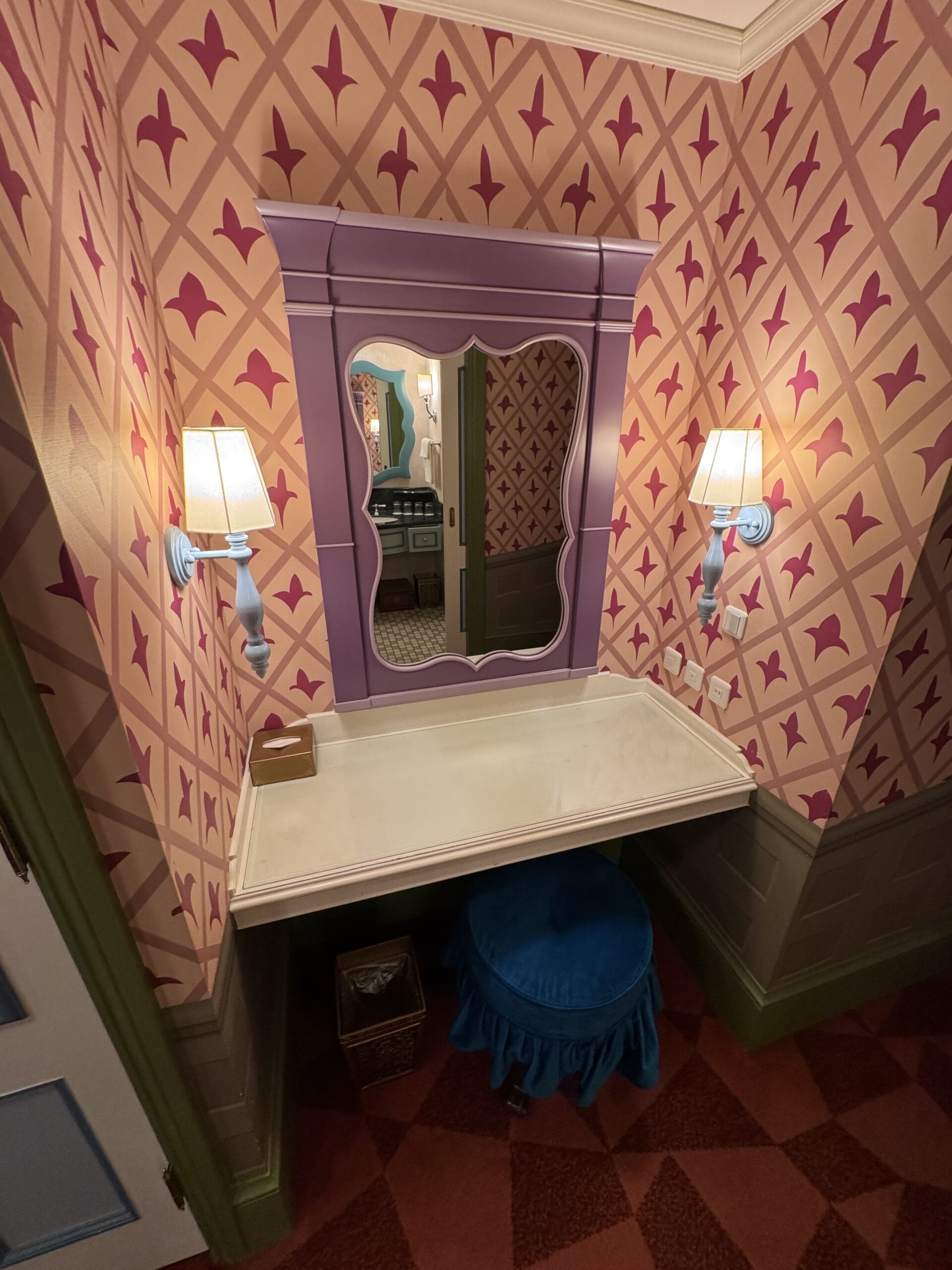 Purple vanity with a decorative mirror evokes an Alice in Wonderland hotel vibe in this whimsical pink patterned room.