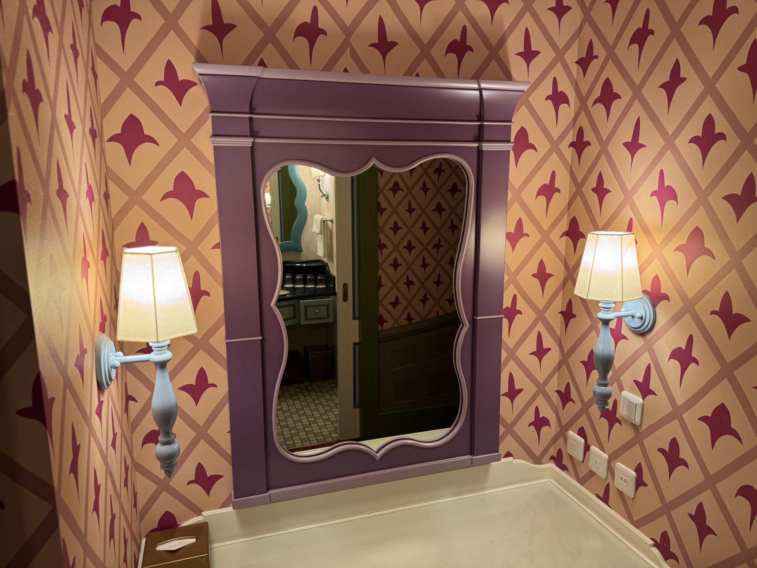 A purple-framed mirror and wall lamps on patterned wallpaper in an Alice in Wonderland hotel room tour scene.