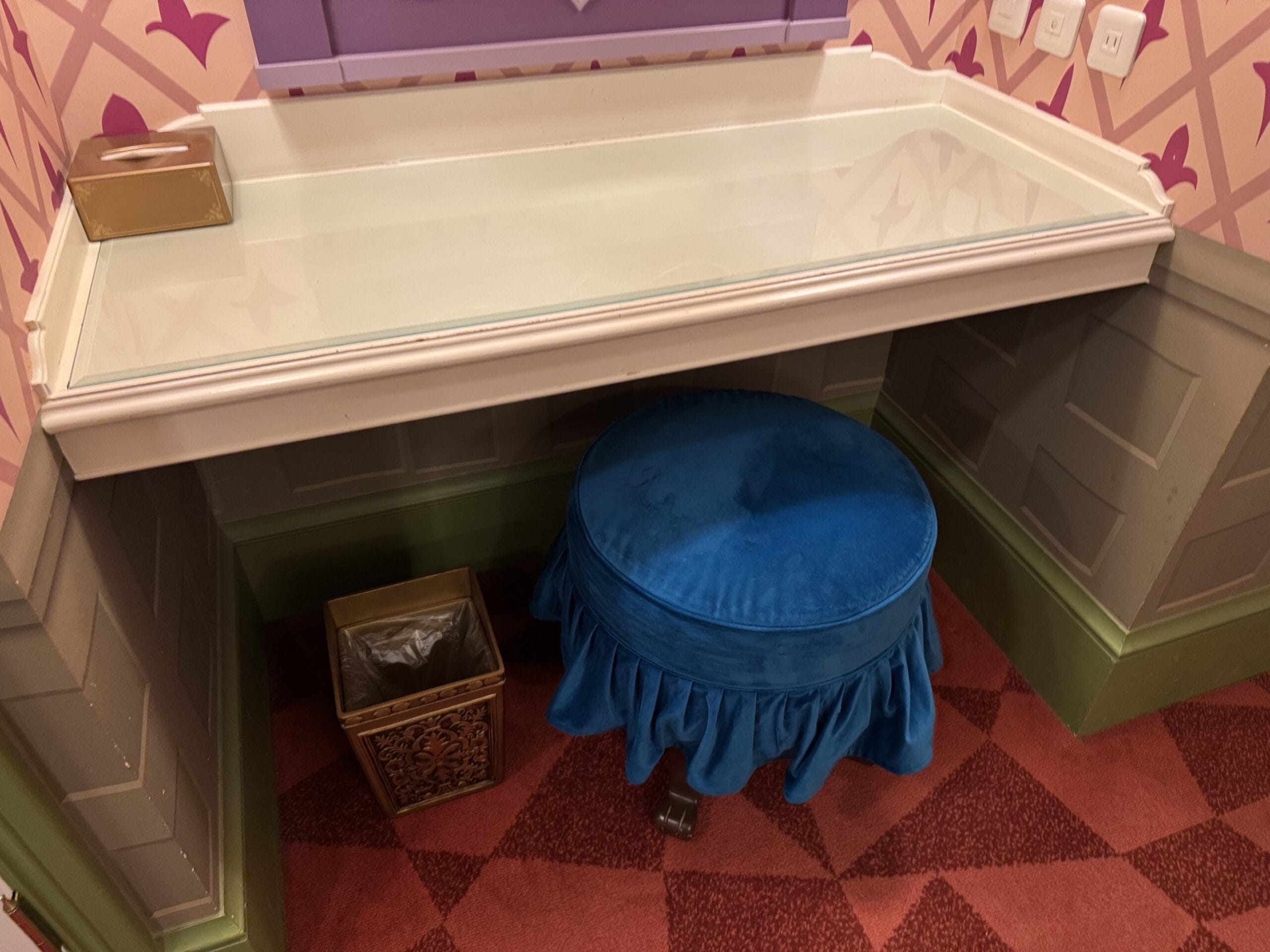A blue cushioned stool and glass-top desk set on a red-patterned carpet in the Alice in Wonderland hotel.