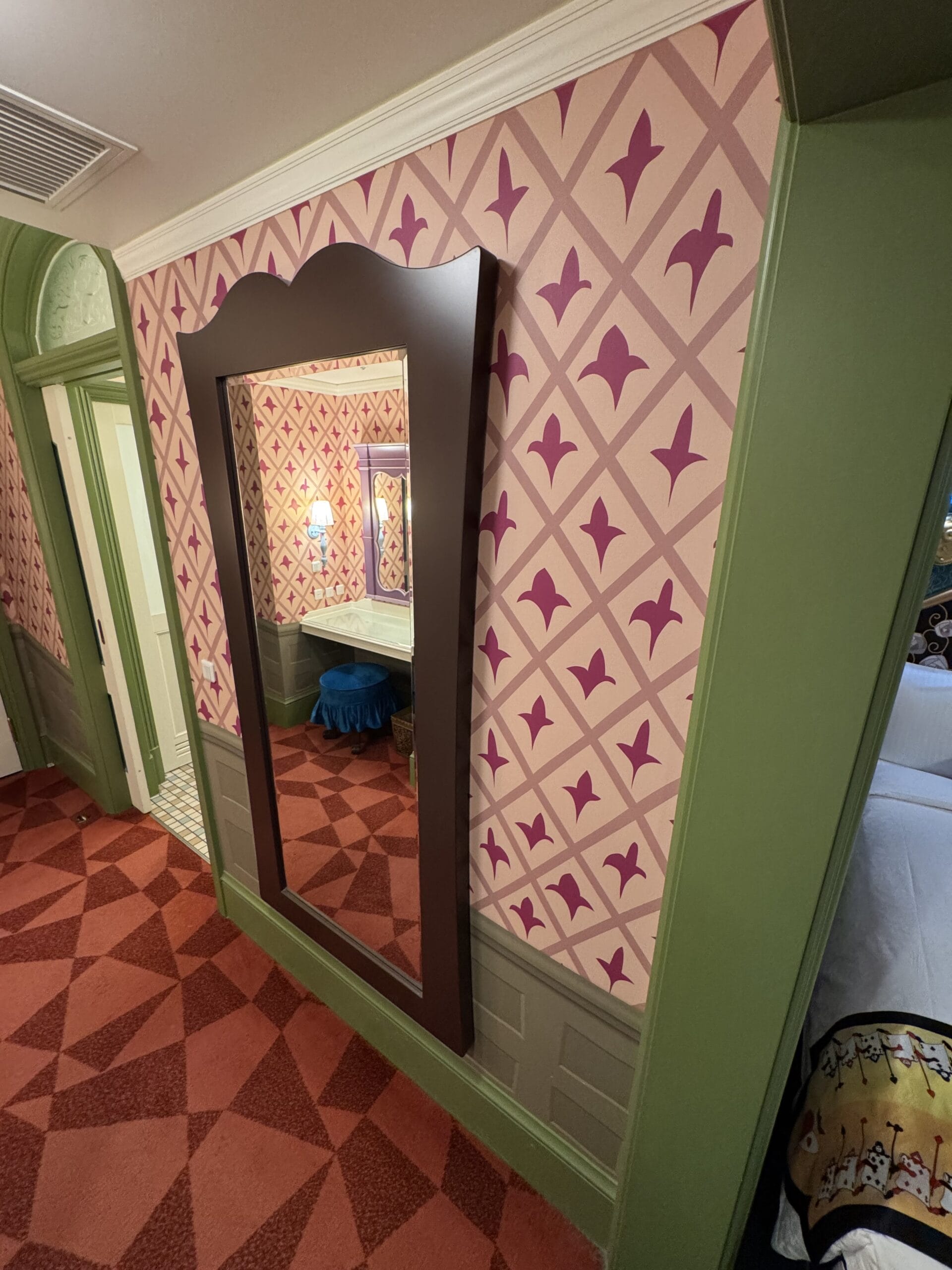 Large framed mirror in a colorful Alice in Wonderland Hotel Room reflects a lamp, blue stool, and bed.