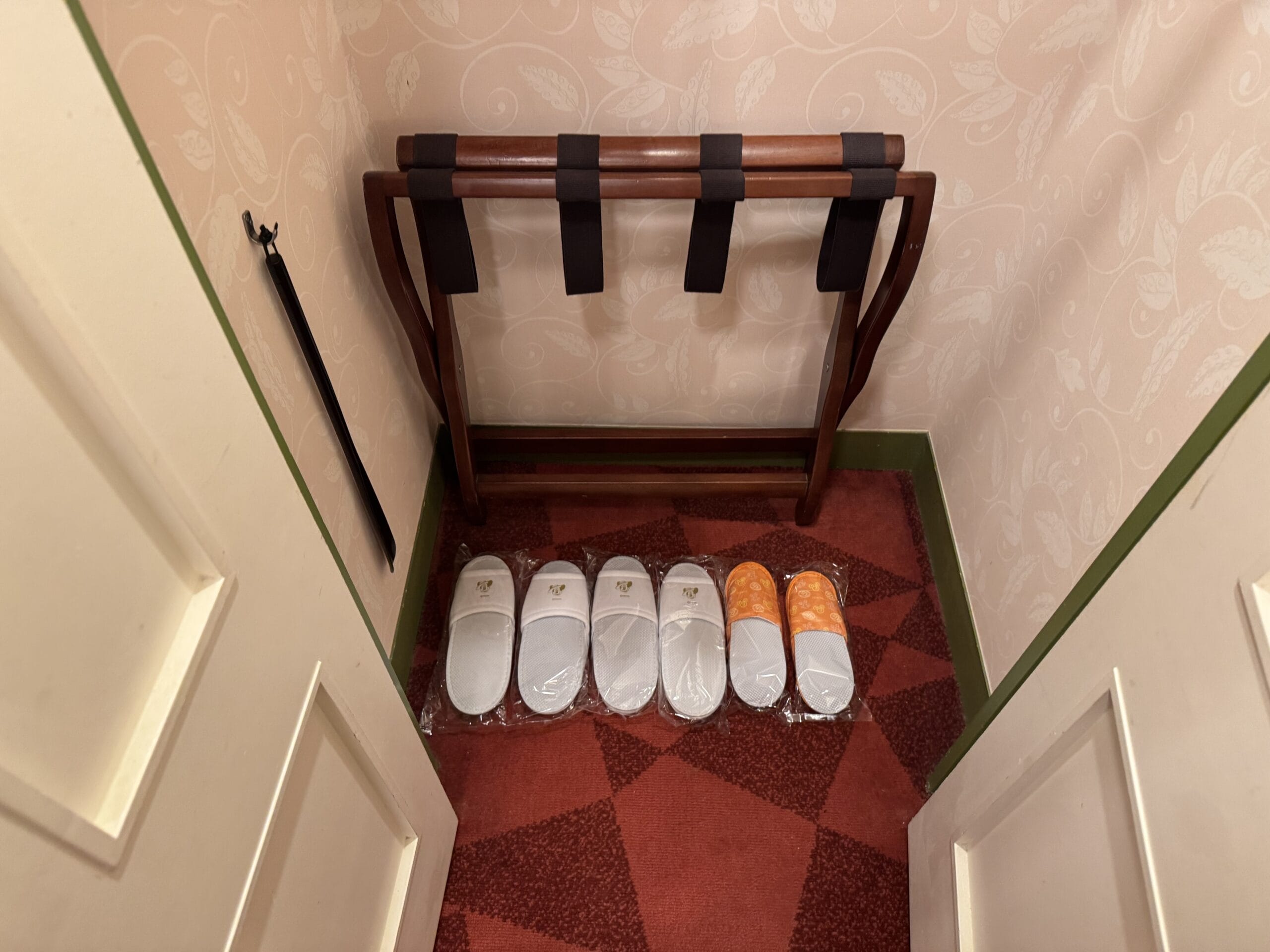 Five pairs of slippers and a wooden luggage rack in a small Tokyo Disneyland Hotel closet with patterned carpet and wallpaper.