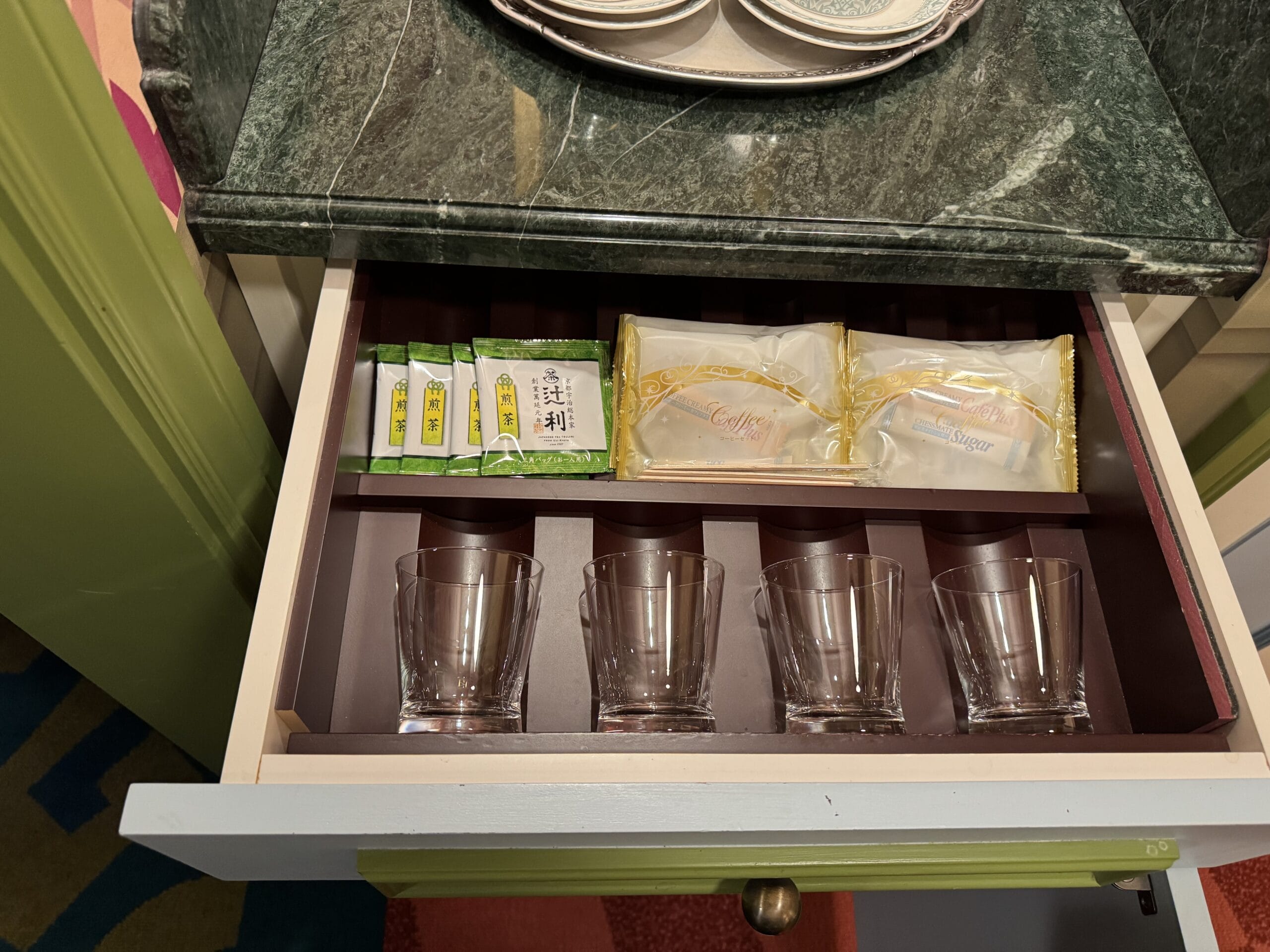 Open drawer in an Alice in Wonderland Hotel Room with glasses, tea, coffee, and sugar on a dark wooden organizer.