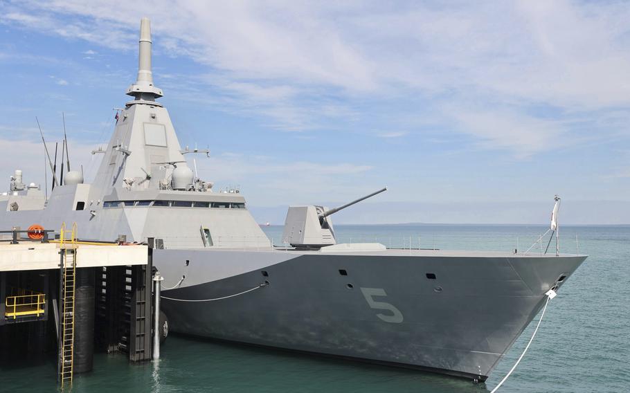 A modern naval frigate with a distinctive angular, stealth design moored at a pier. The ship has a gray hull with the number “5” visible on its bow, along with a prominent pyramidal superstructure and various radar and communication arrays. A gun turret is visible on the forward deck. 