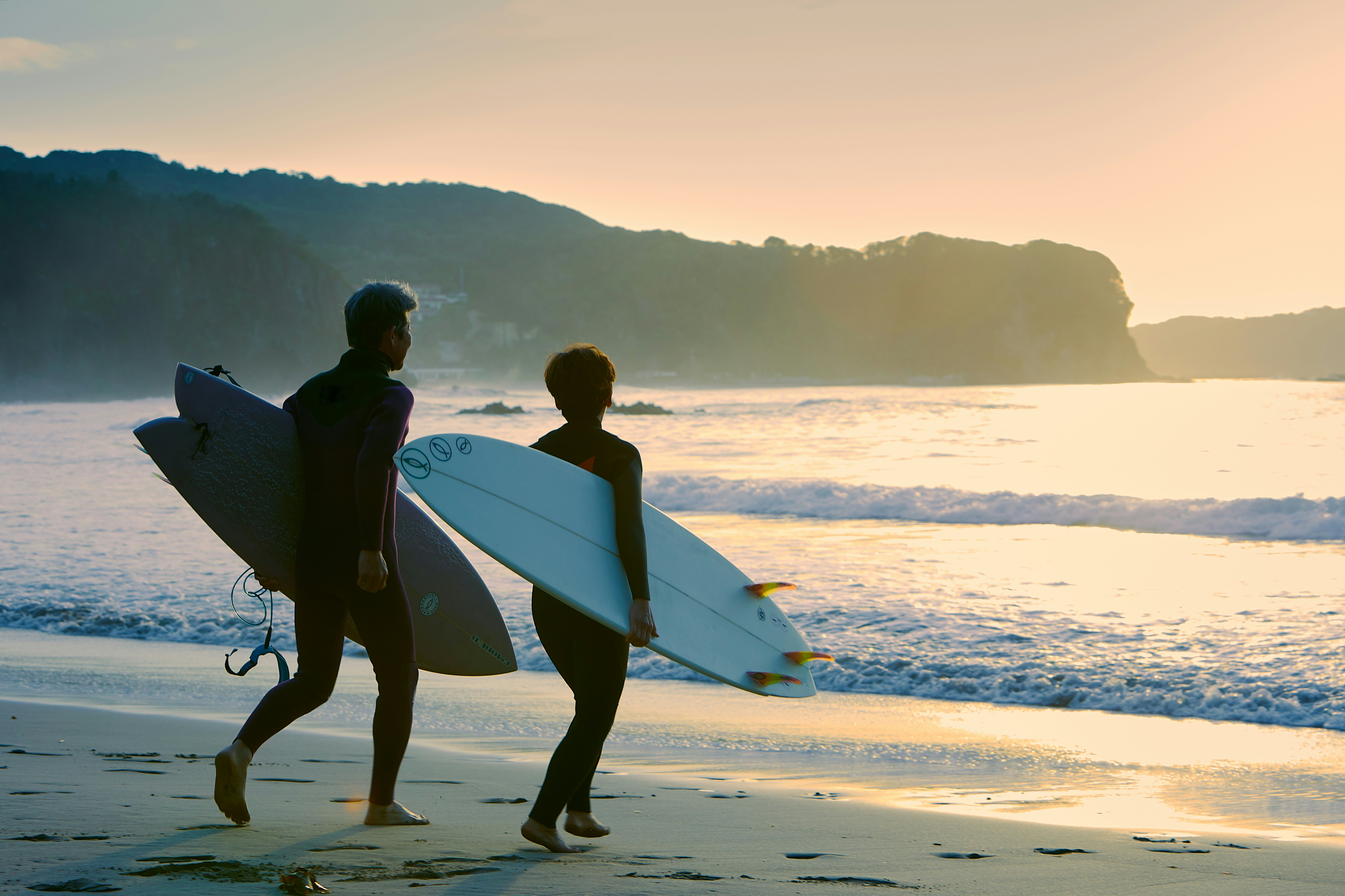A man and woman head into the sea with their surfboards under their arms as the sun sets.