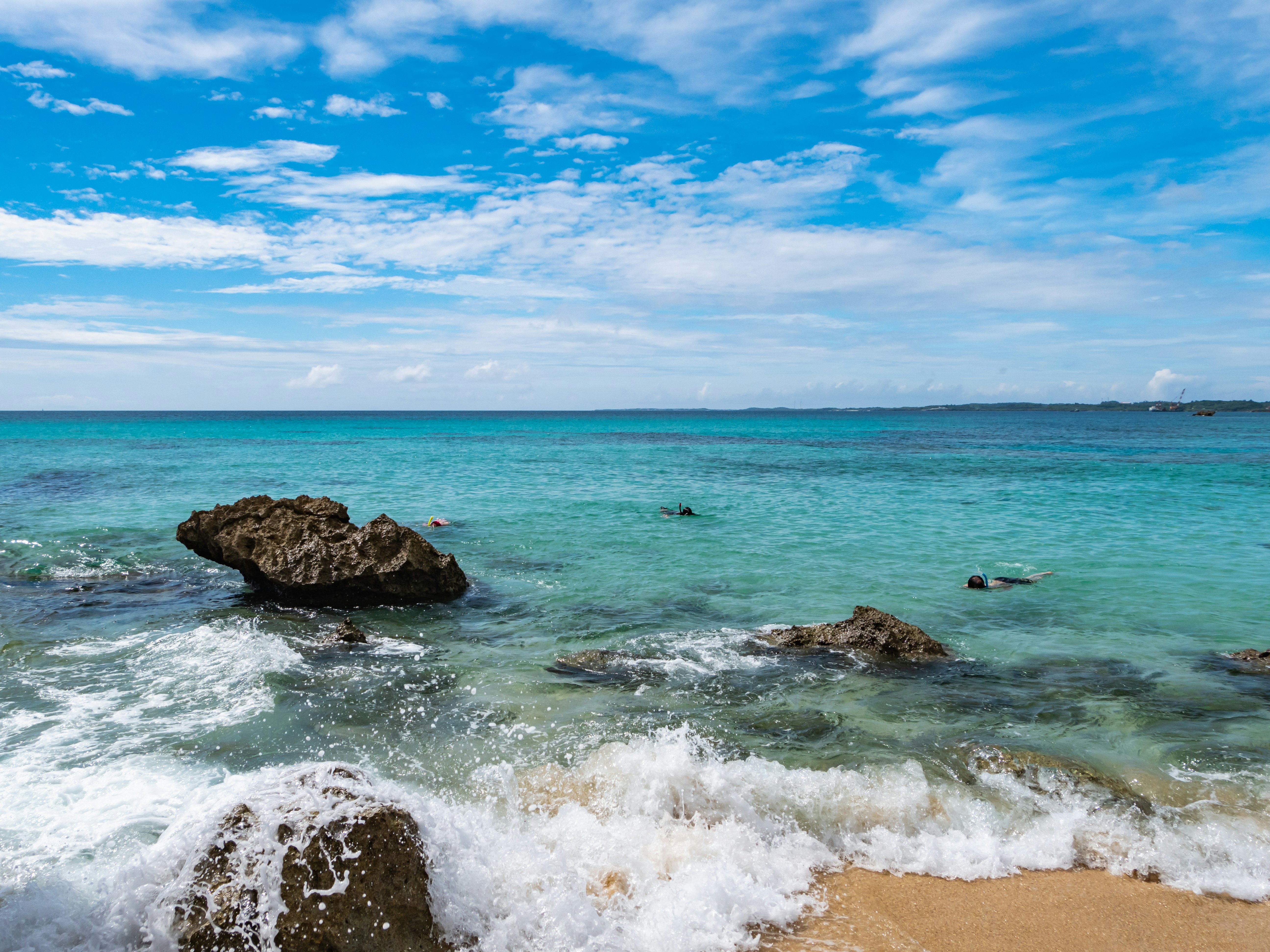 People snorkeling in bright blue ocean water with large rocks in the foreground on a sunny day.