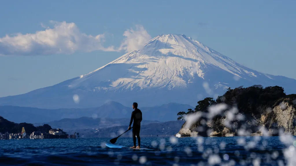 A man paddles a "puddle" board while waiting for waves with Mount Fuji in the background near Zaimokuza Beach in Kamakura, Japan, January 10, 2025.