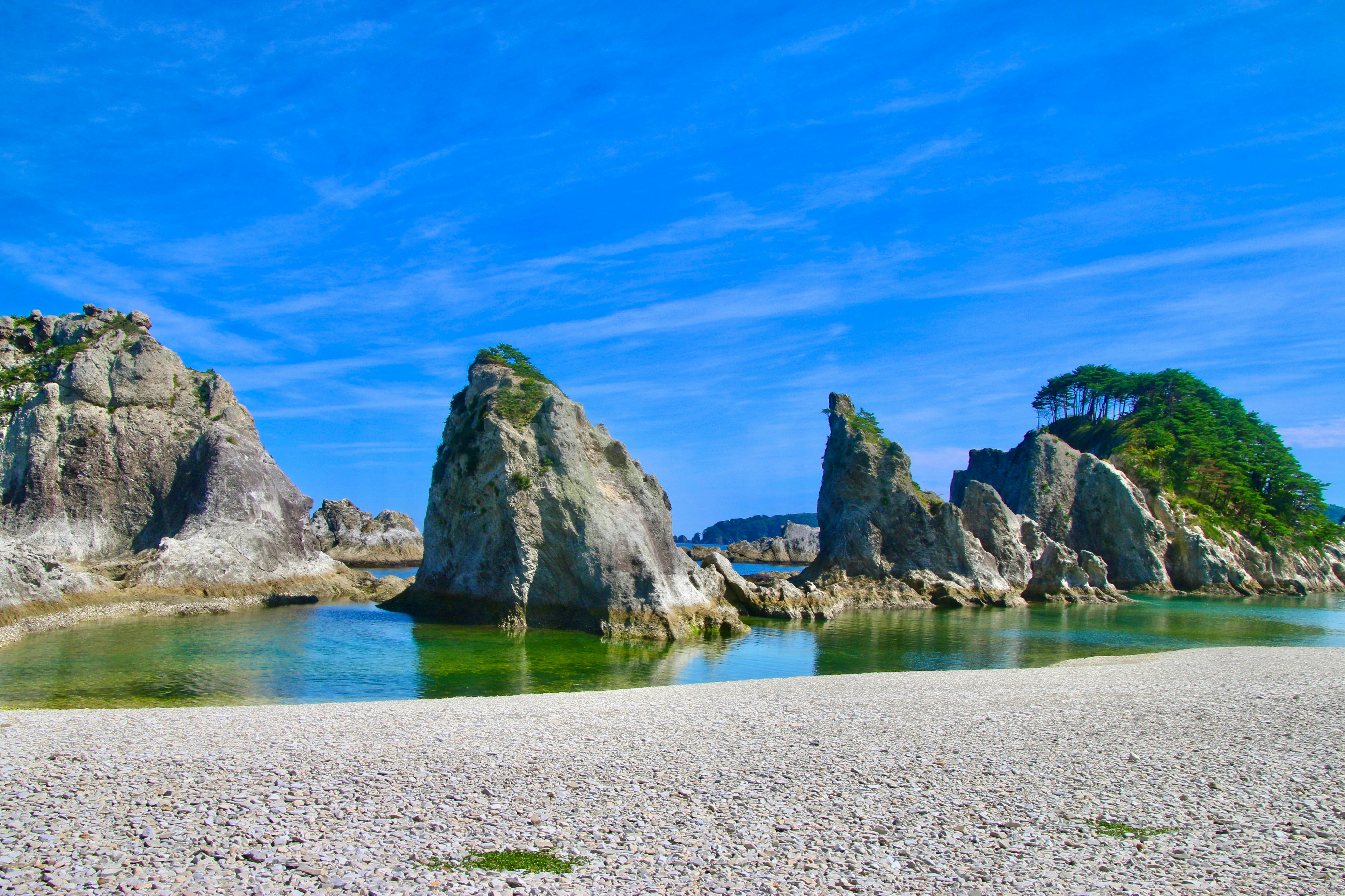 Large sea stacks in shallow water beside a beach of smaller rocks on a sunny day.