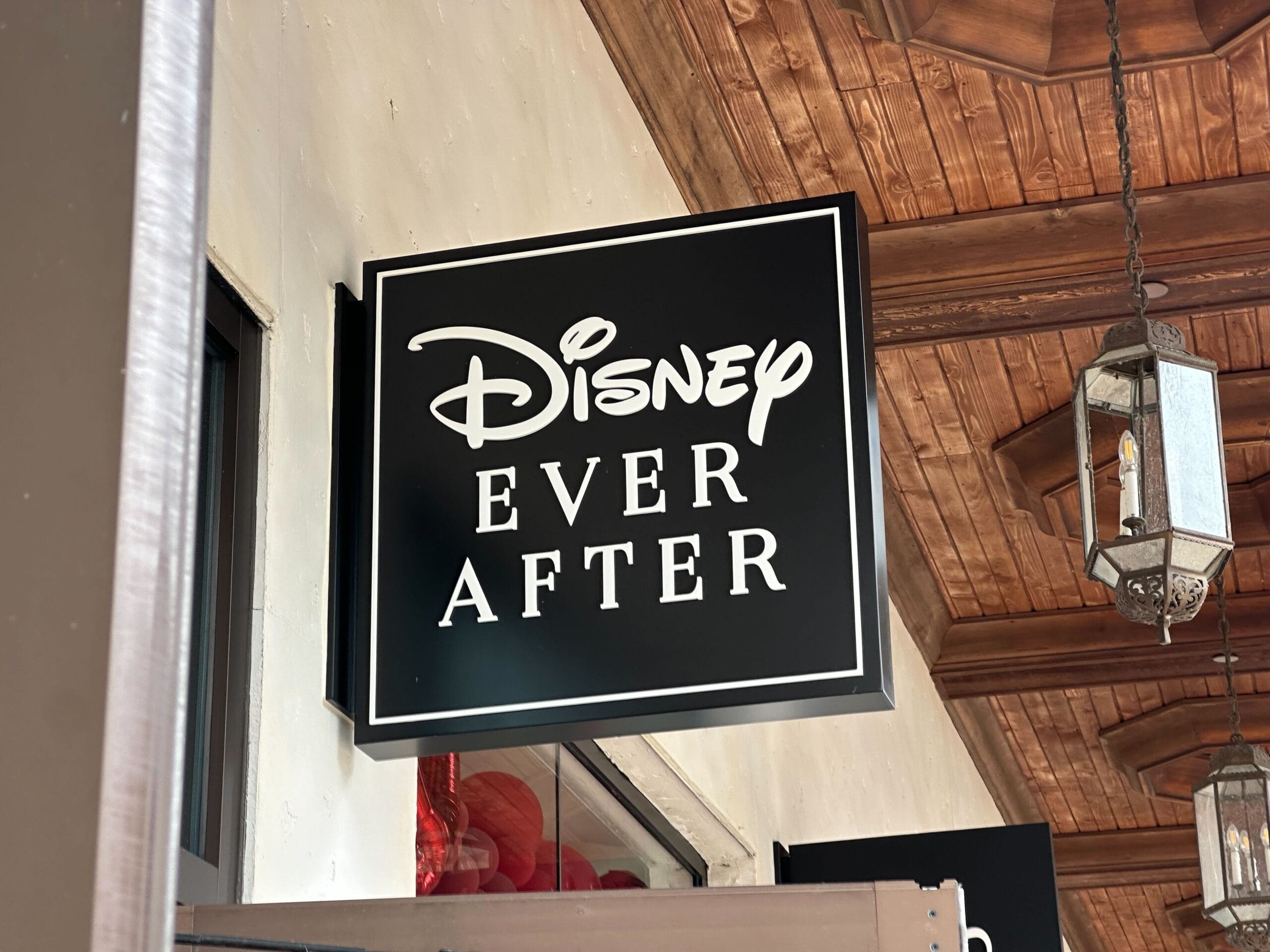 A wall-mounted black sign, part of the charming Disney Springs decor, reads "Disney Ever After" in white letters beneath a wooden ceiling adorned with hanging lanterns.