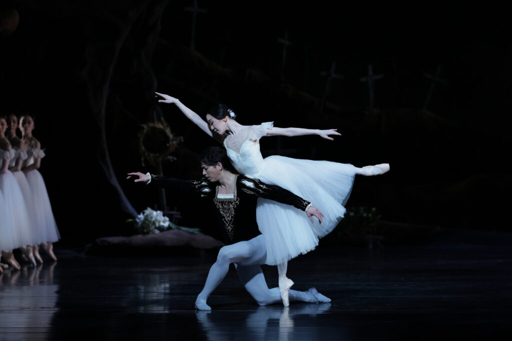 Two dancers perform a pas de deux onstage, the man kneeling as his partner does an arabesque on pointe and leans over him, resting against his back. She wears a long white Romantic tutu and he wears gray tights and a dark velvet tunic.