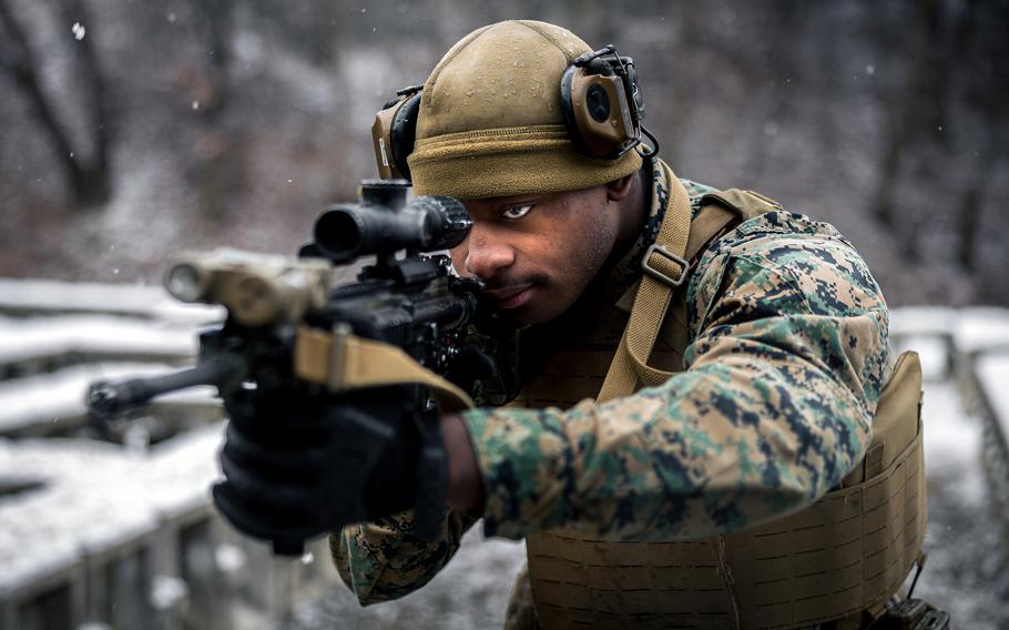 A marine in camouflage uniform and tactical gear aims a rifle with an optical scope in a snowy outdoor environment. The person is wearing a tan helmet with mounted equipment, protective eyewear and a tactical vest.