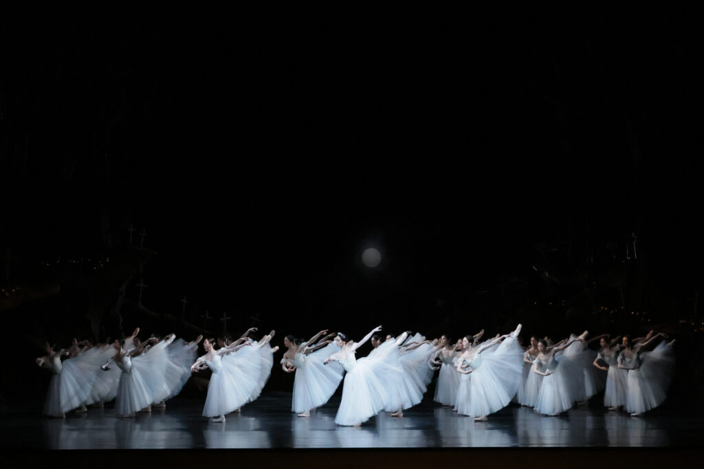 Onstage, a large group of corps dancers in neat lines wear long white Romantic tutus. They fondu in arabesque, arms in third, with a slight lean forward.