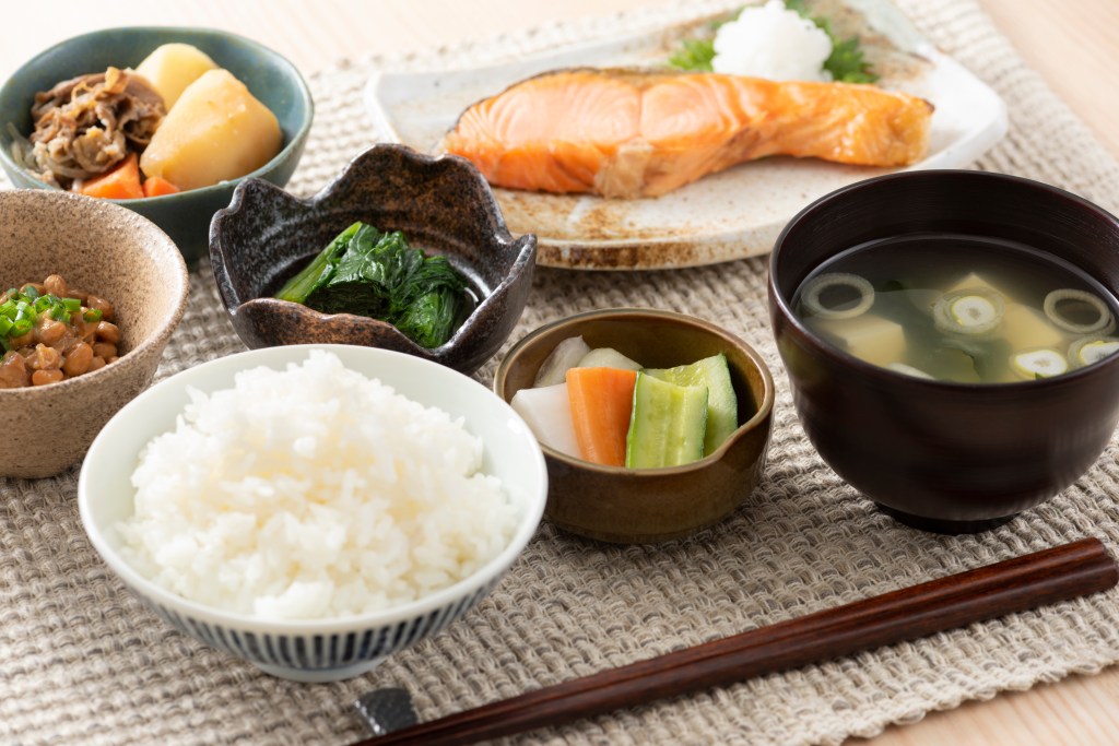 Japanese breakfast spread with rice, miso soup, grilled salmon, and side dishes.