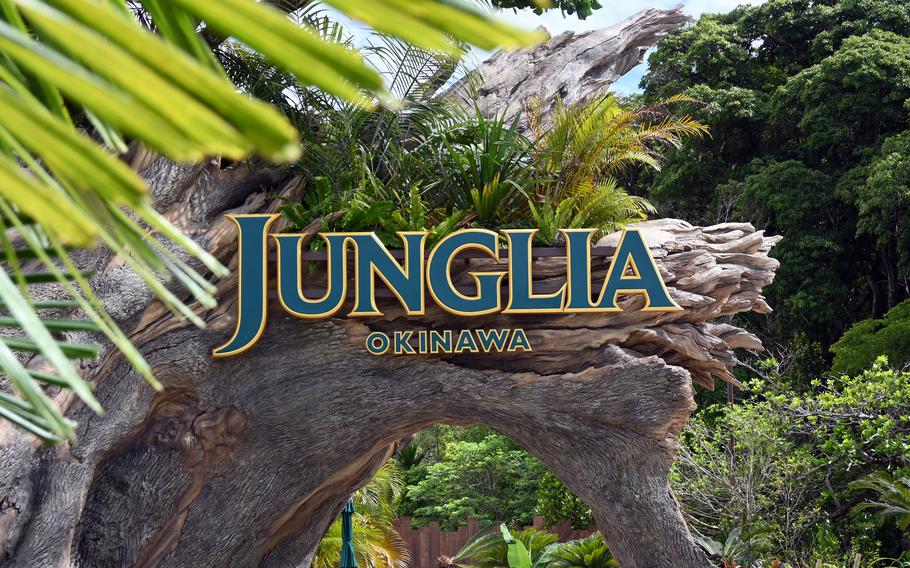 The entrance sign to “Junglia Okinawa” in gold lettering, positioned on a natural rock archway surrounded by tropical vegetation including palm fronds and dense green foliage.