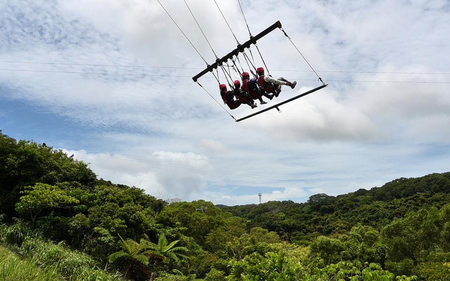 A group of people wearing red helmets are on a swing high above a dense green forest canopy, with rolling hills and a communications tower visible in the distance.