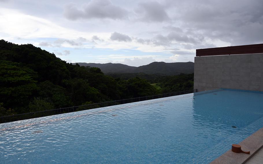 An infinity pool with clear blue water overlooking a lush, forested mountain landscape under a cloudy sky, with modern concrete architecture visible on the right side.