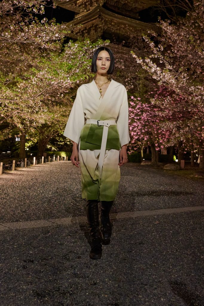 A model wears a green and white belted ensemble with black boots from Dior’s Fall 2025 collection, walking under illuminated cherry blossoms at night in Kyoto.