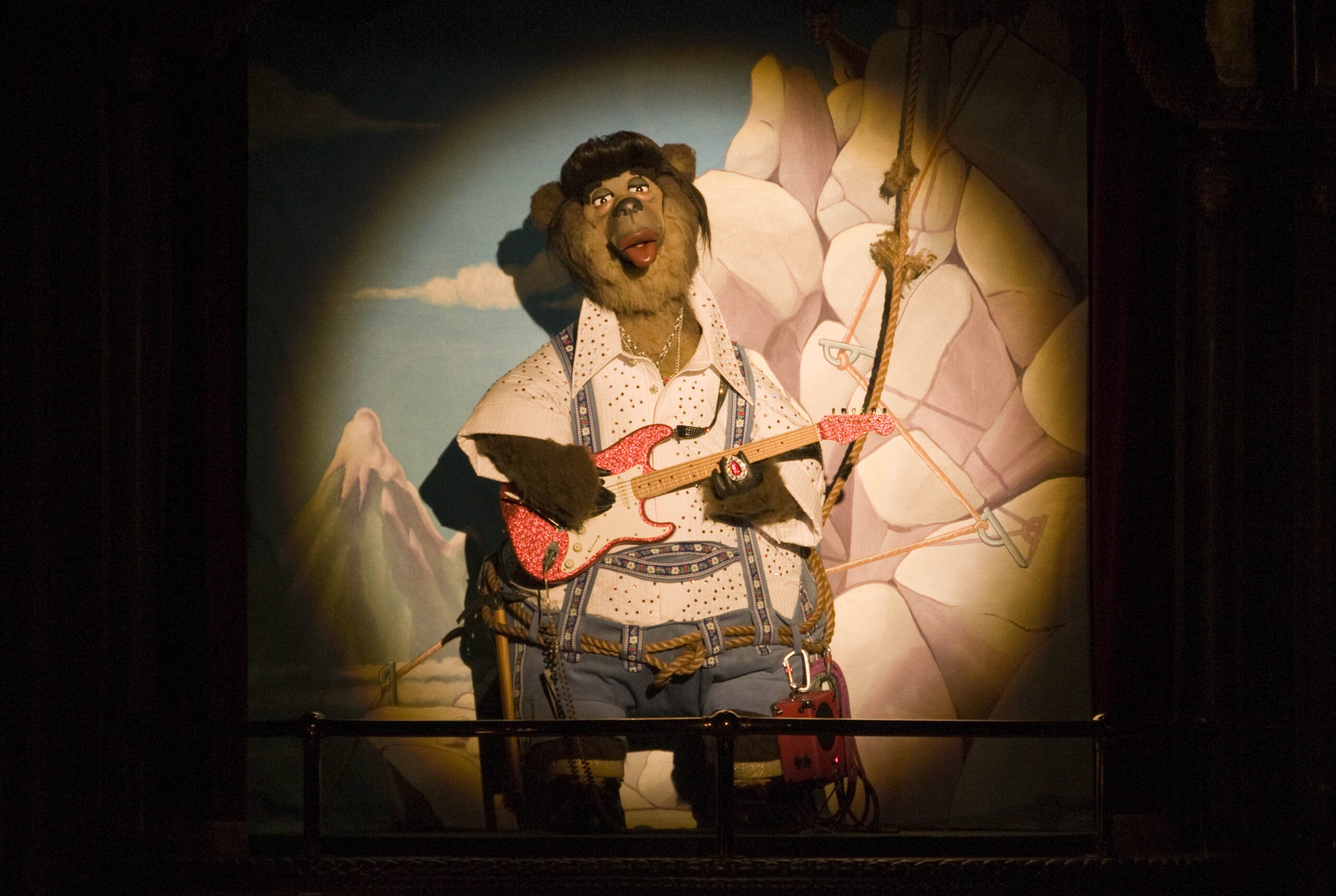 An animatronic bear wearing western clothes plays a pink guitar on stage with a mountain backdrop.