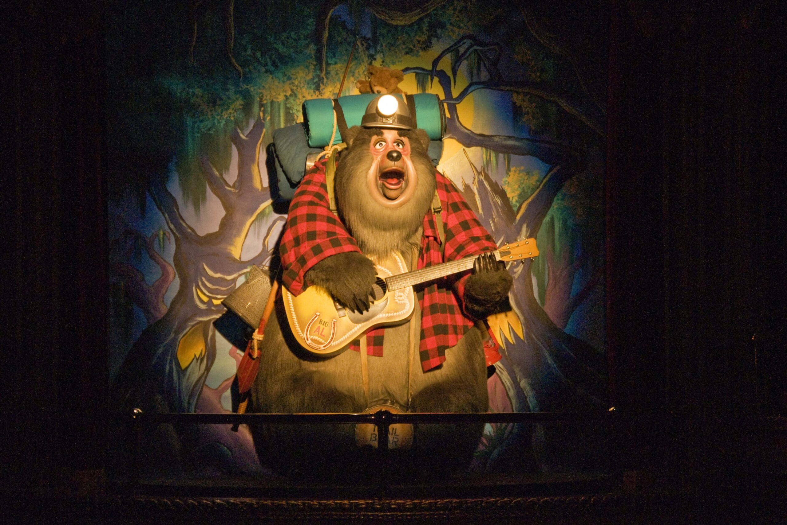 An animatronic bear in a plaid shirt and hat plays a guitar on stage with a forest backdrop.