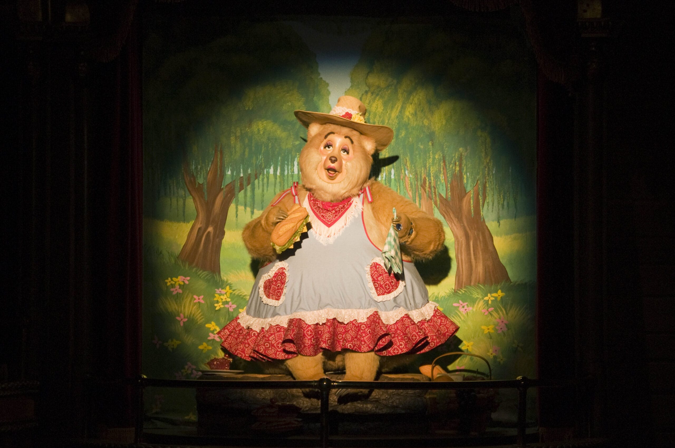 A bear in a dress and hat stands on stage, singing, with a painted forest backdrop behind her.
