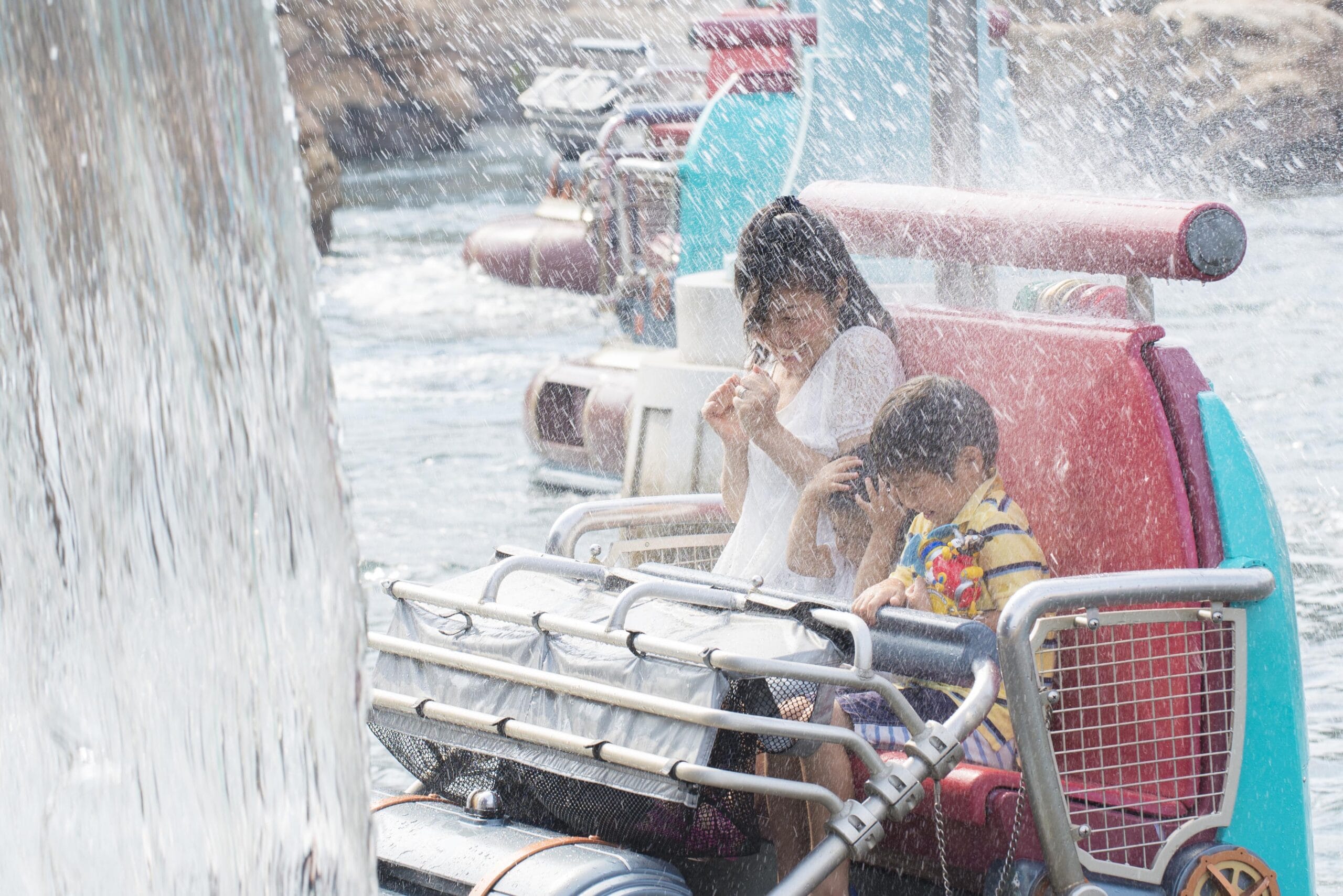 Two children on a theme park water ride get splashed with water, shielding their faces and laughing in the spray.
