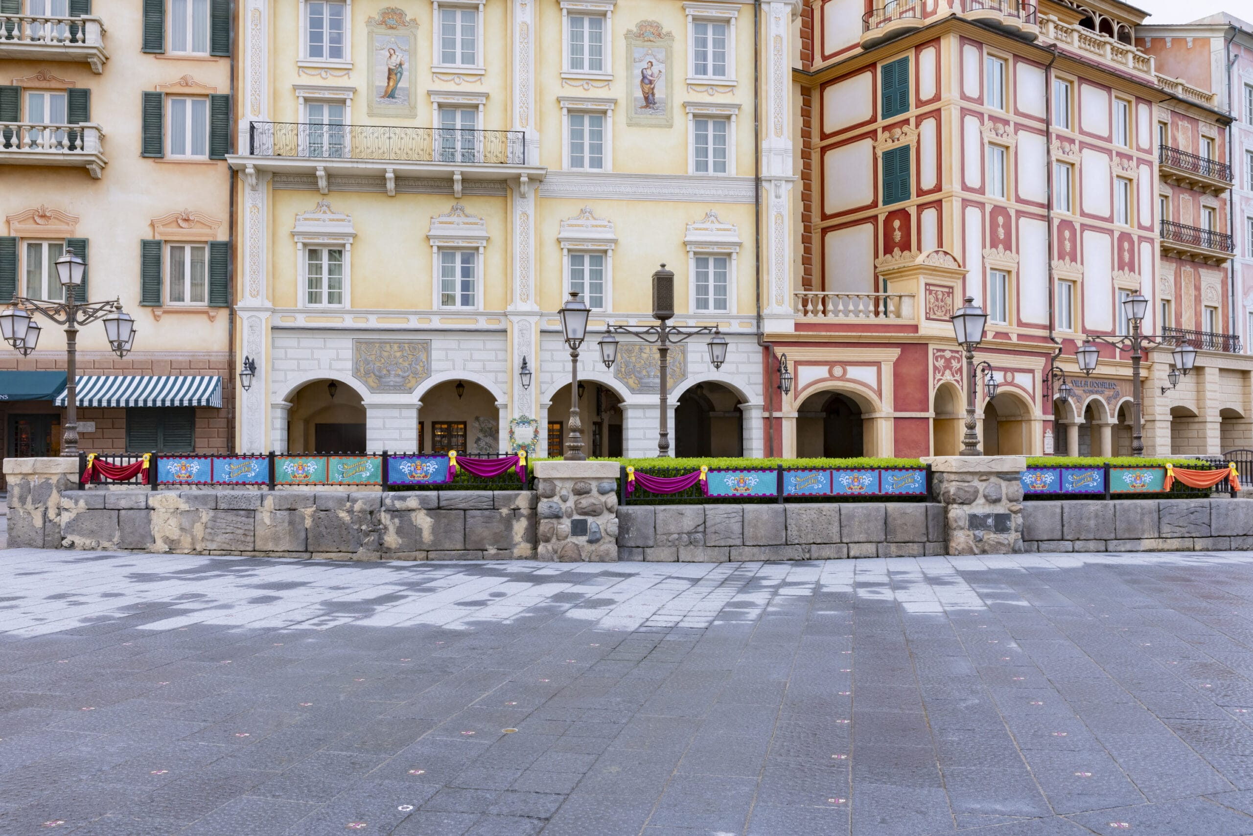 A colorful, decorated fence stands in front of ornate European-style buildings on an empty, stone-paved plaza.