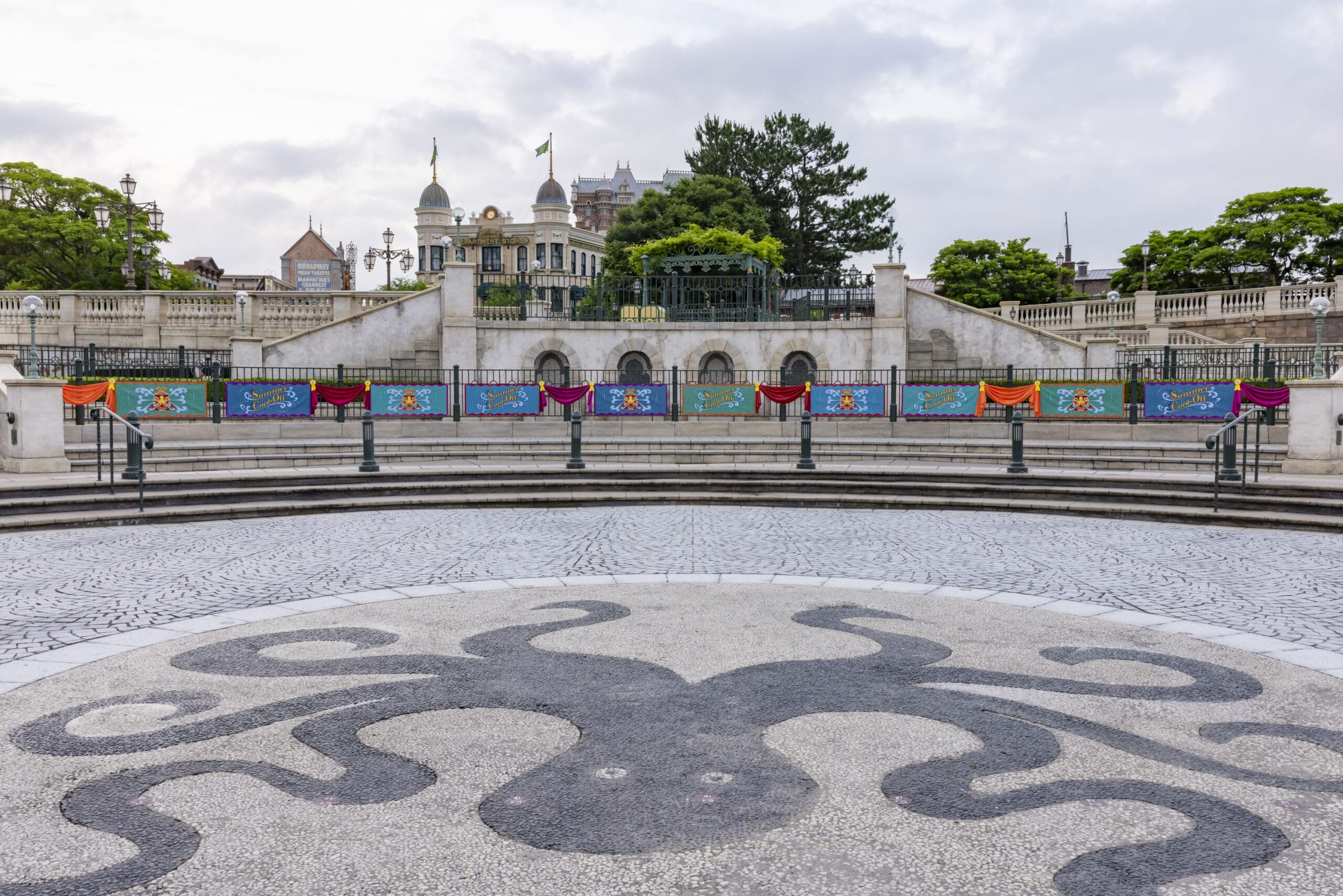 Circular plaza with an octopus design on the ground, colorful banners, and a grand building in the background.