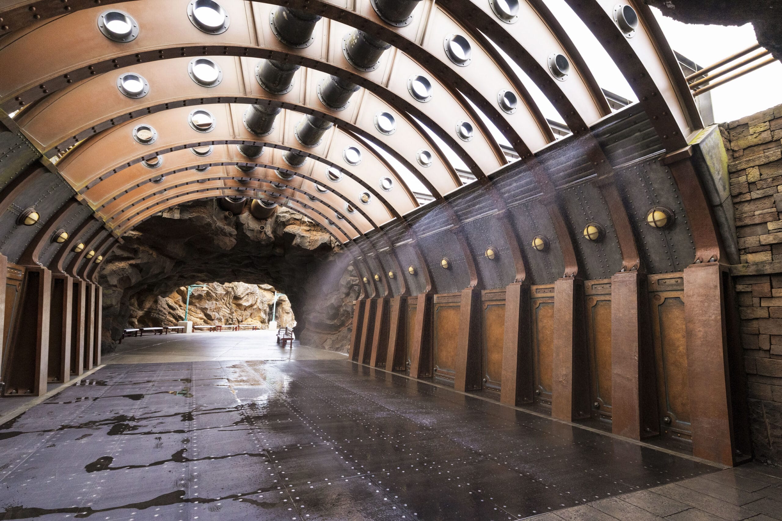 A person with a stroller walks through a steampunk-style tunnel with metal rivets and circular windows.