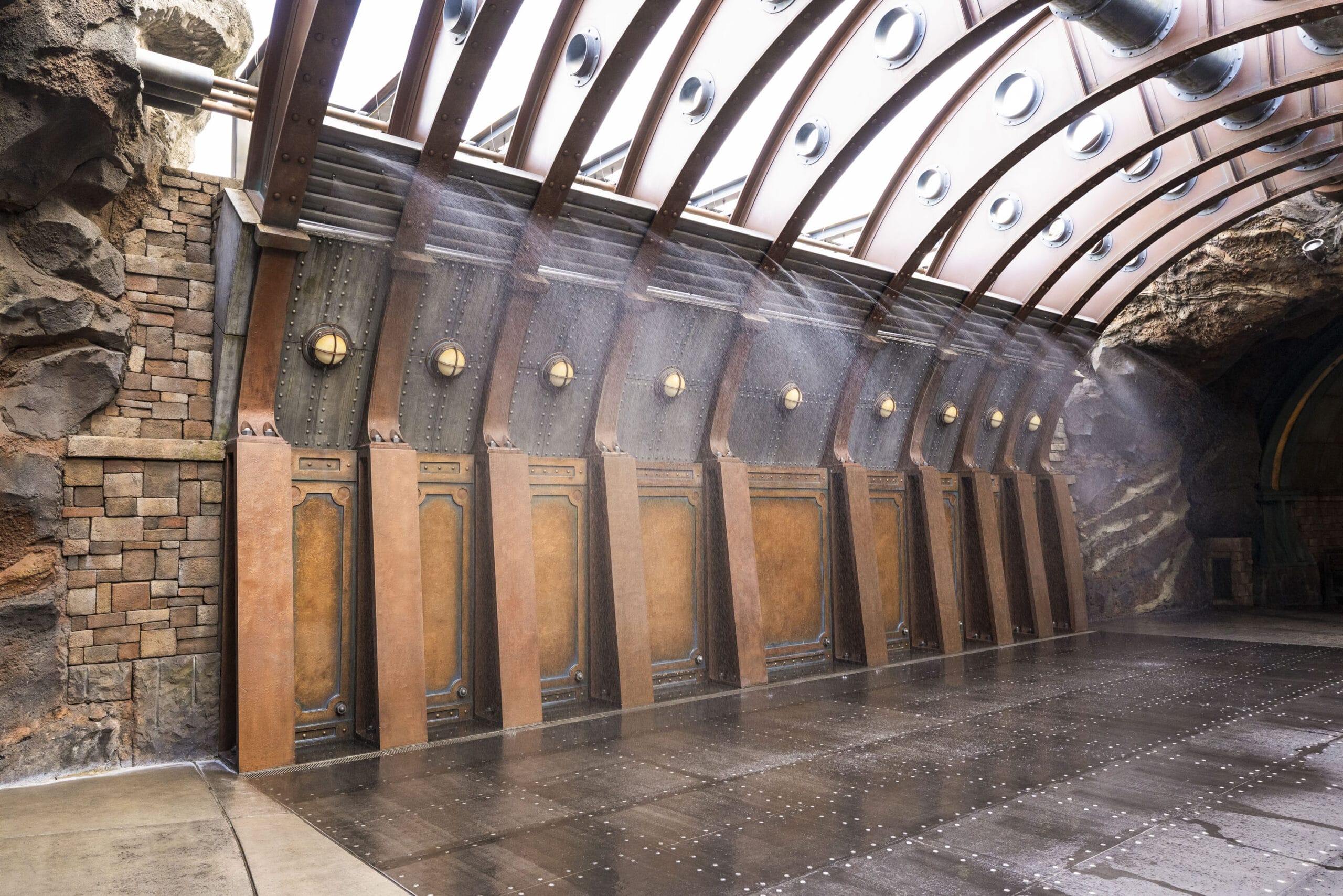 Steampunk-inspired corridor with metal and glass ceiling, mist spraying, and rock and metal walls.
