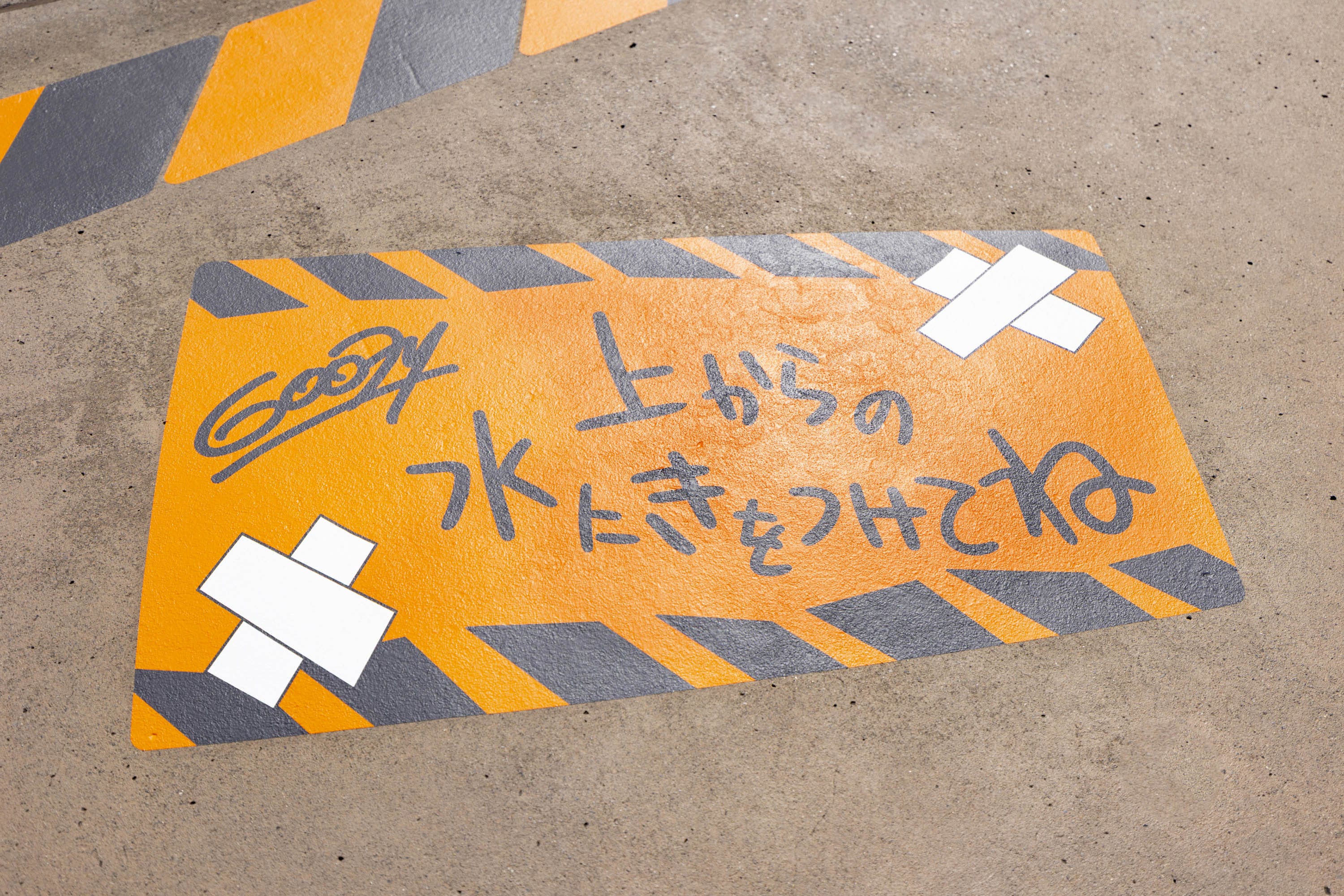 Yellow caution sign with Japanese text and black stripes on concrete, decorated with white tape illustrations.